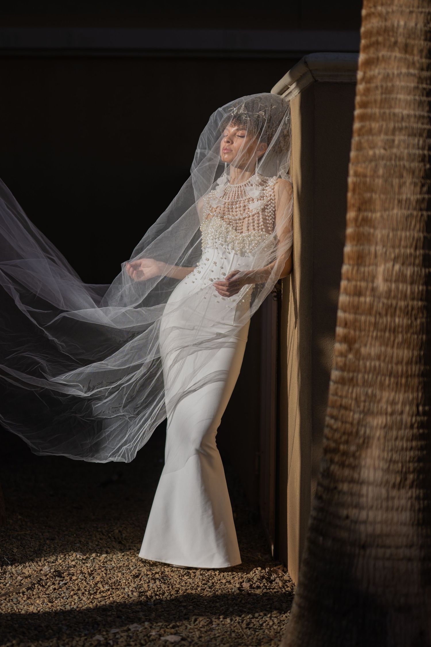 Bride in fitted white mermaid gown with lace bodice and flowing veil, posed against wooden post in dramatic lighting.