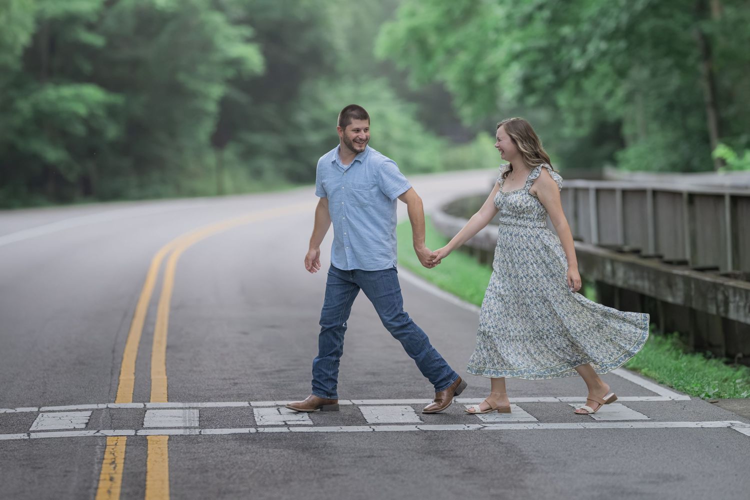 Couple holding hands crossing a road surrounded by lush green trees, woman in floral dress, man in blue shirt and jeans.