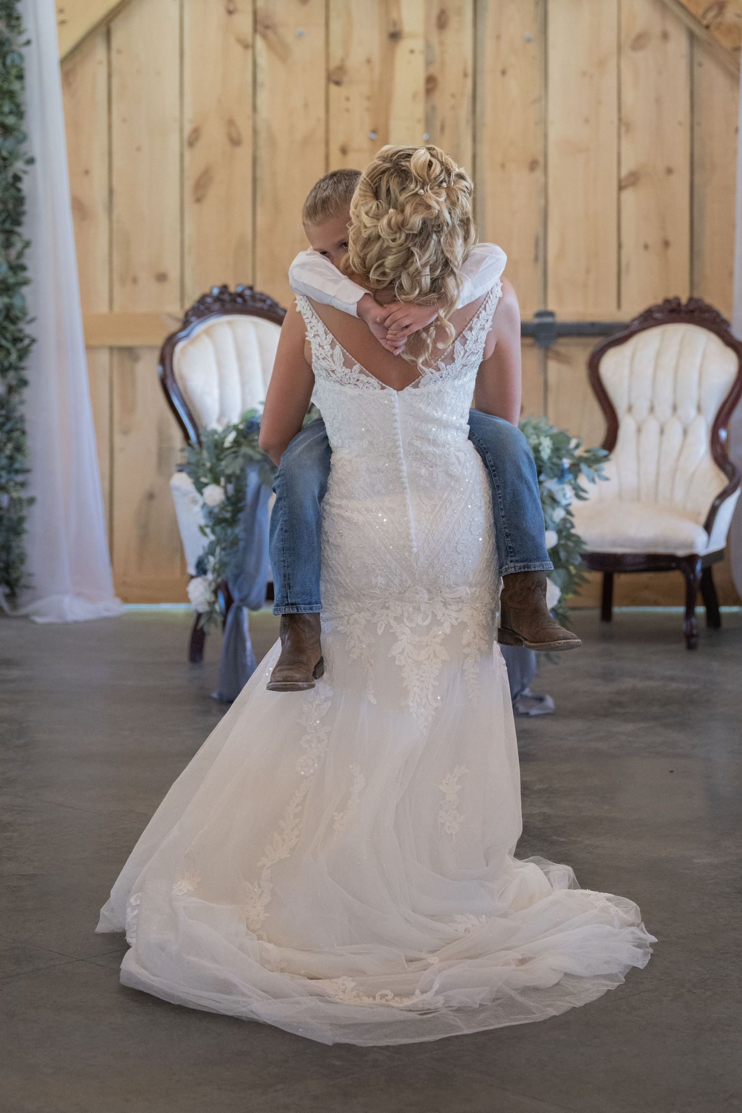 Bride in lace mermaid wedding gown carrying a child in jeans and cowboy boots inside a rustic barn venue.