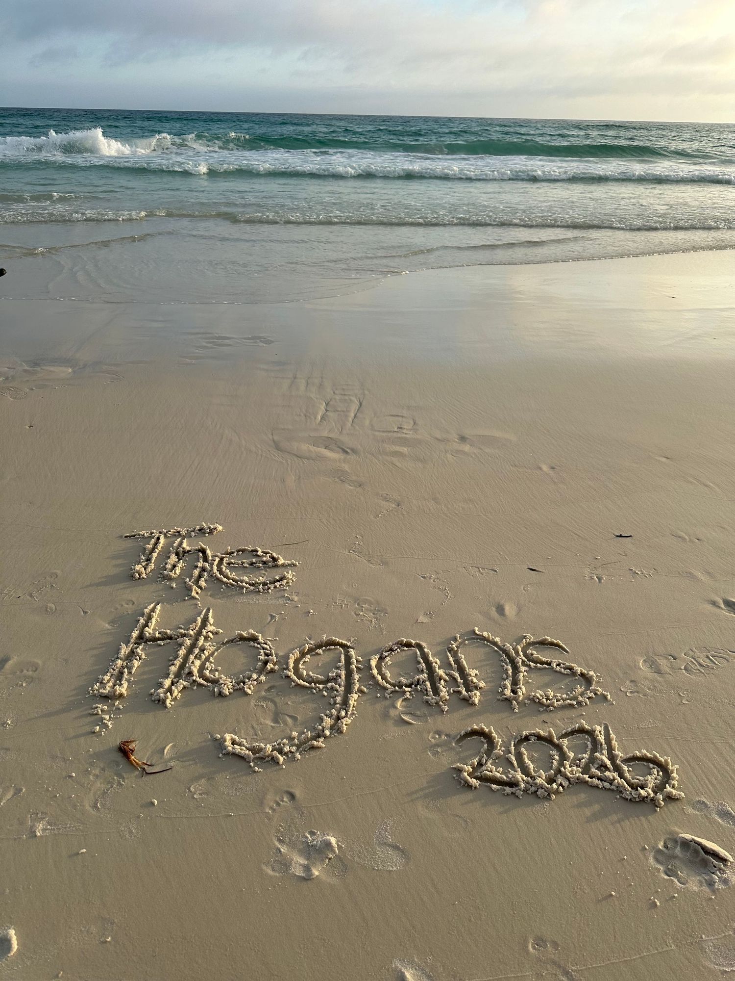 The Hogans 2026 written in sand on a beautiful beach with turquoise ocean waves in the background.