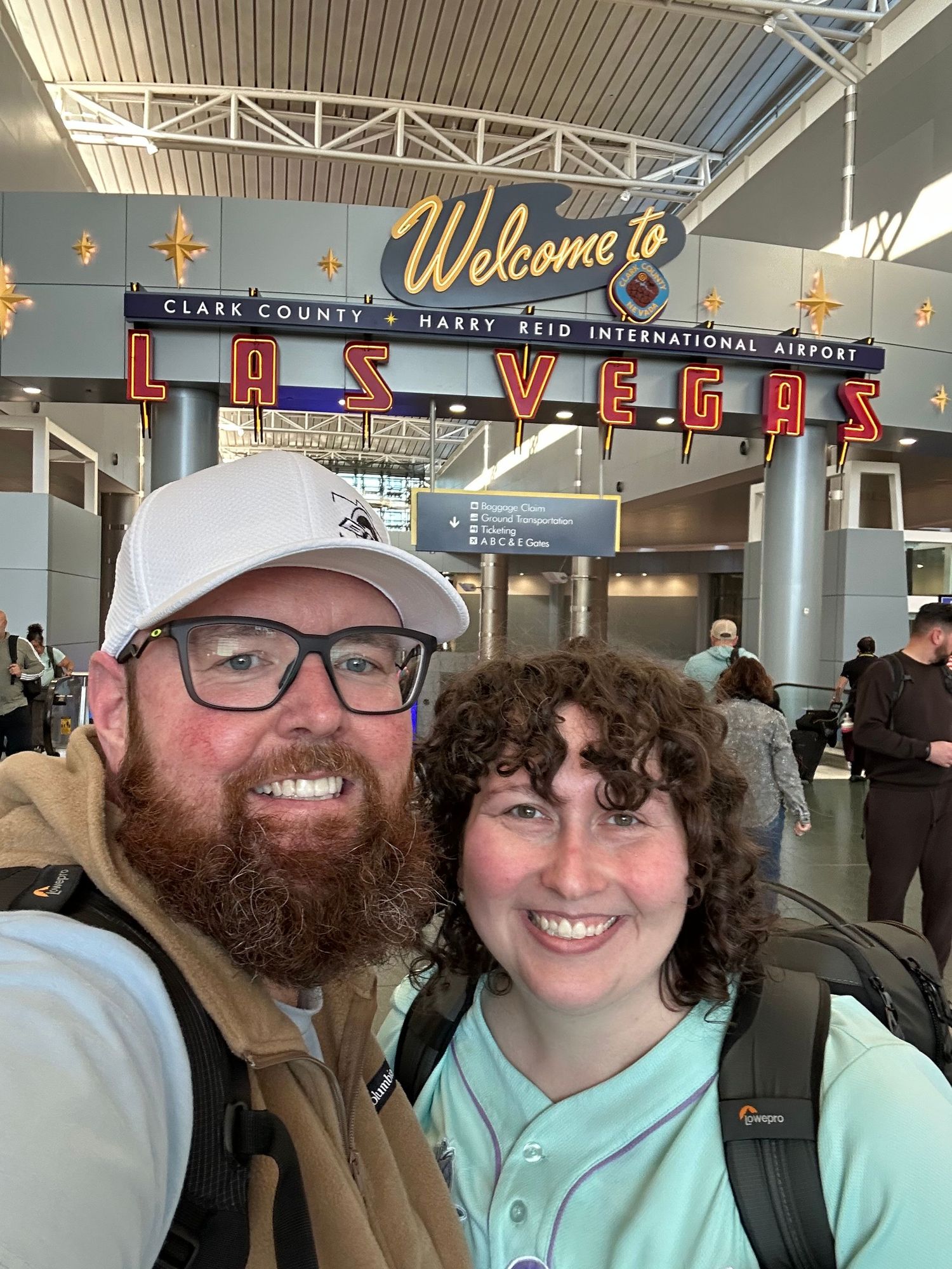 Couple smiling for selfie in front of Welcome to Las Vegas Harry Reid International Airport sign.