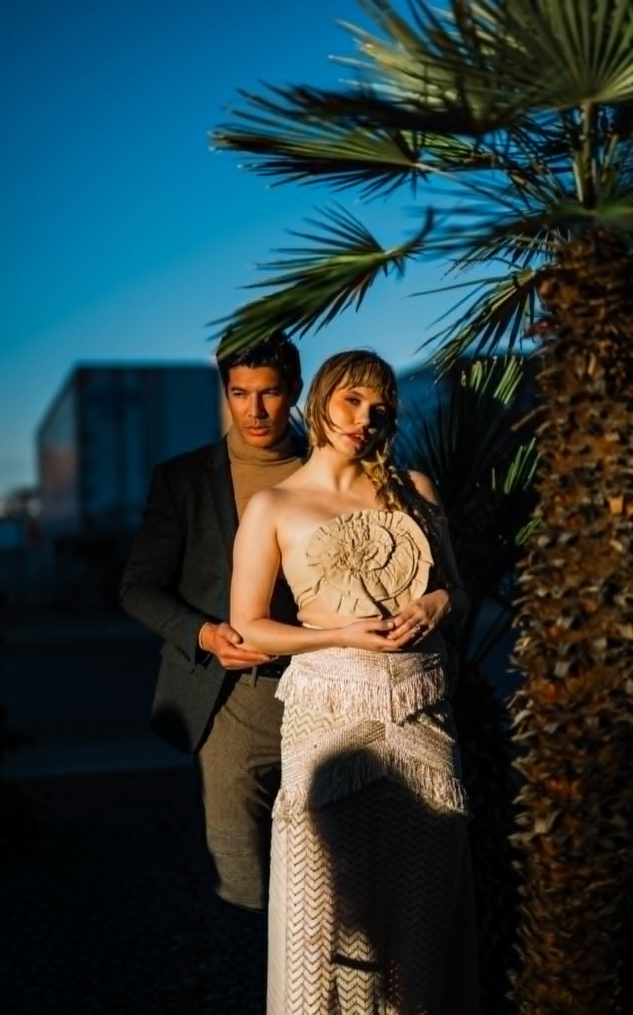 Couple posing near a palm tree at golden hour, woman in elegant fringed dress, man in dark suit, blue sky backdrop.