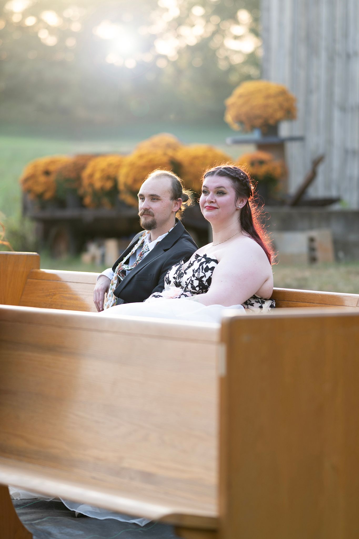 Couple sitting on wooden church pew outdoors at fall wedding with golden mums and barn backdrop.