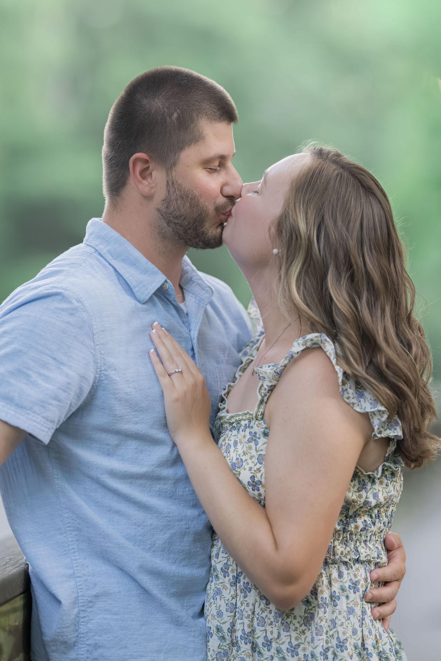 A couple shares a romantic kiss outdoors, woman in floral dress and man in blue shirt embracing in soft natural light.