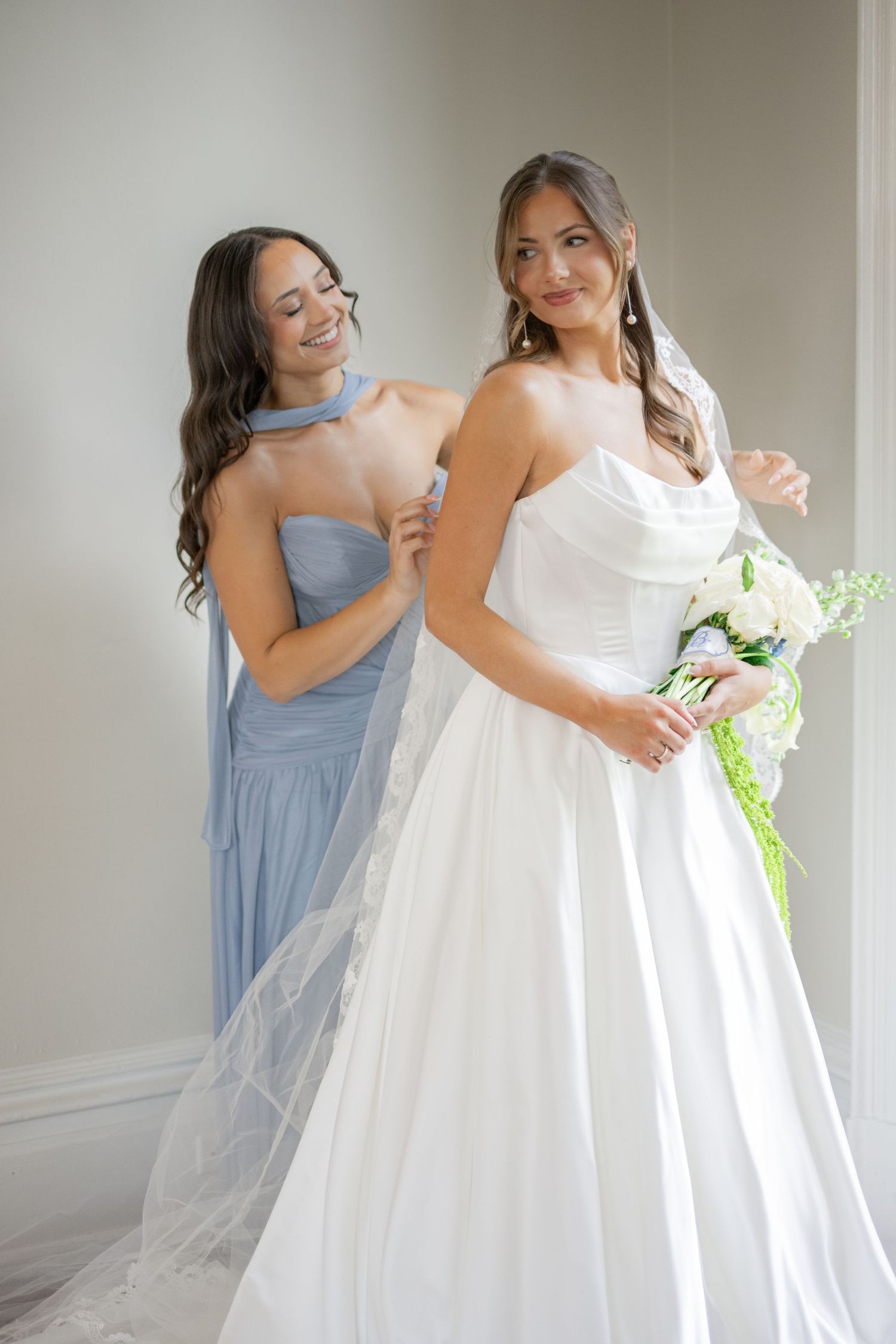 Bride in white strapless ball gown holding bouquet with bridesmaid in blue dress helping her prepare.
