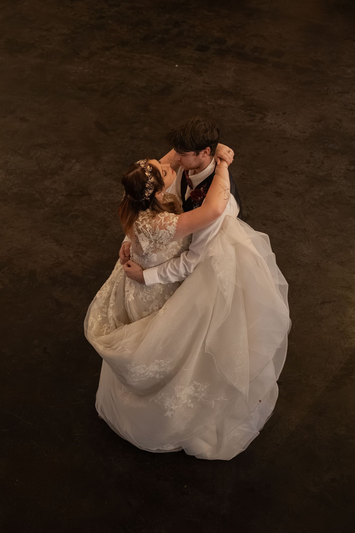 Bride and groom sharing first dance, aerial view of white lace wedding gown flowing on dark floor.