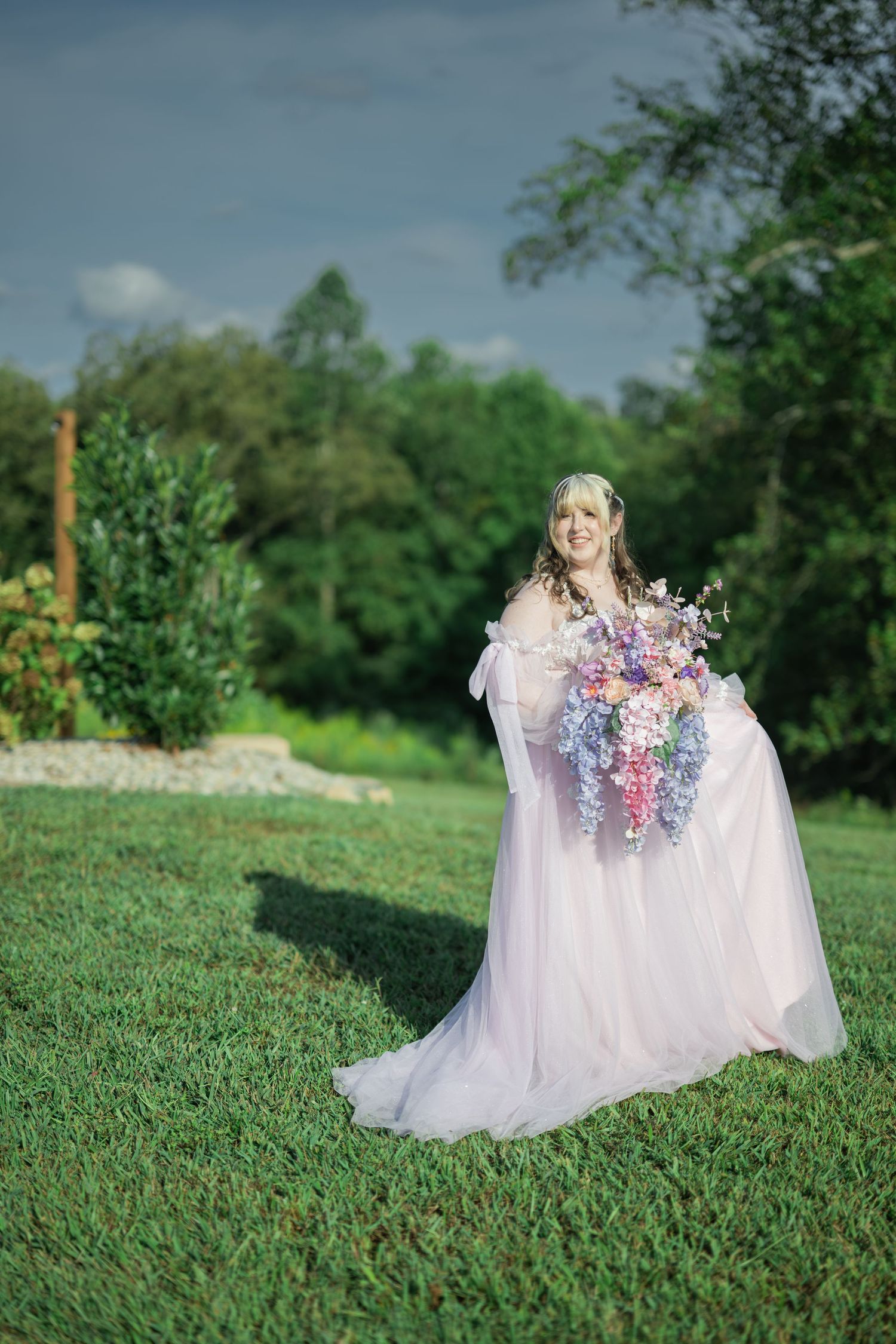Bride in flowing pink gown holds colorful bouquet on green lawn with trees and cloudy sky backdrop.