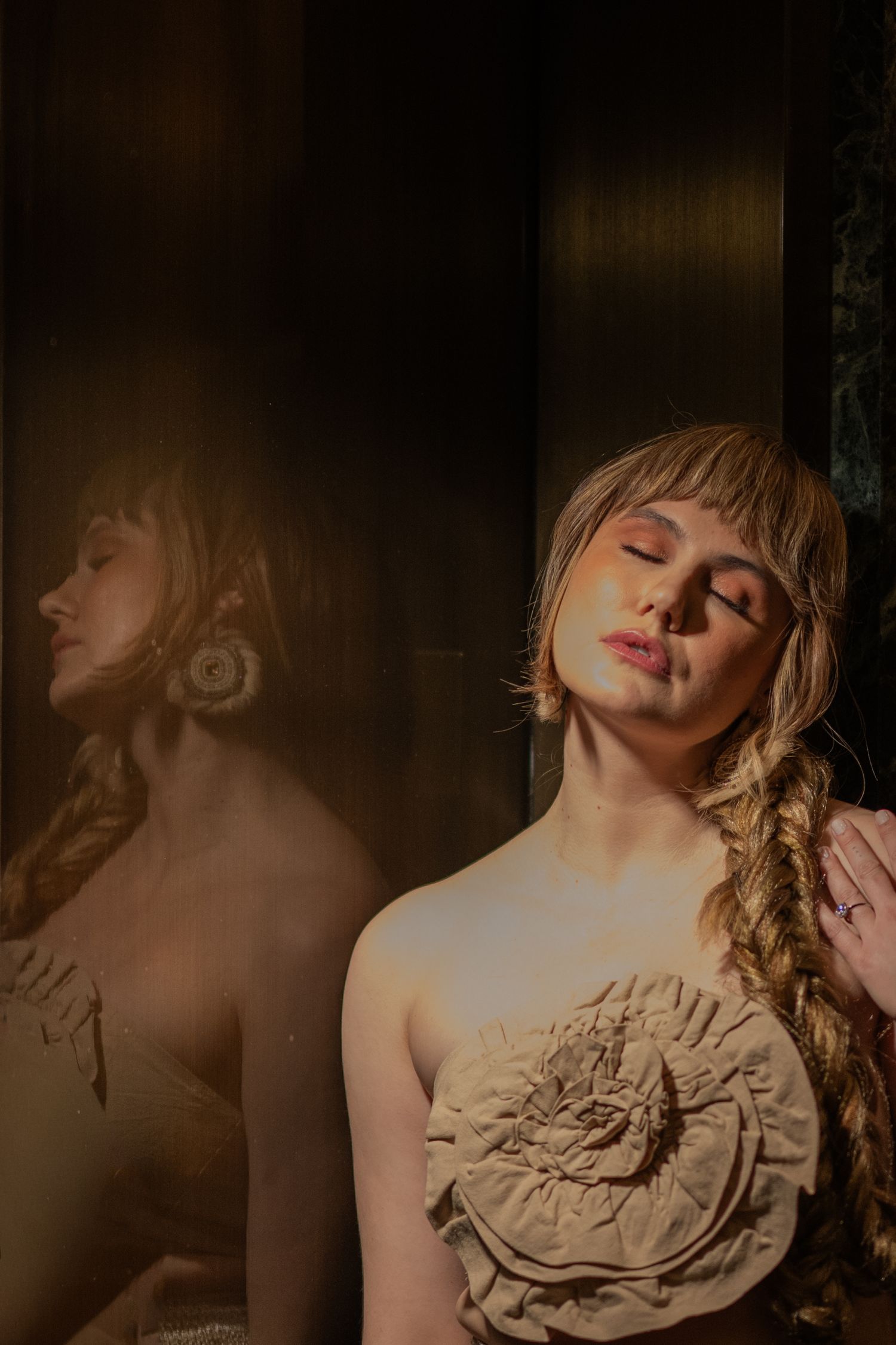 A woman with blonde braided hair and a floral top poses with eyes closed beside a dark mirror reflection.