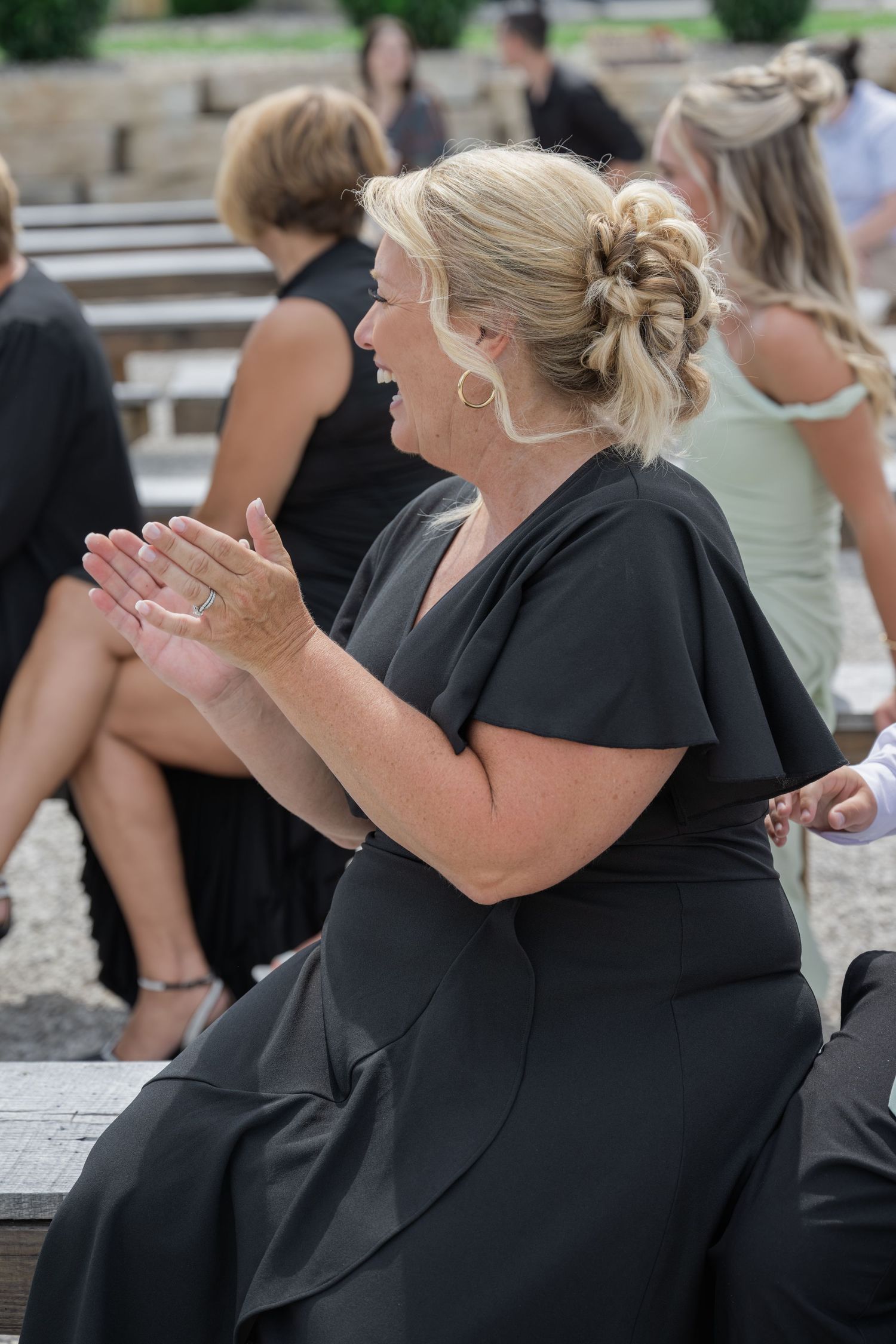 Woman in black dress clapping and laughing at outdoor event, blonde updo hairstyle, gold hoop earrings.