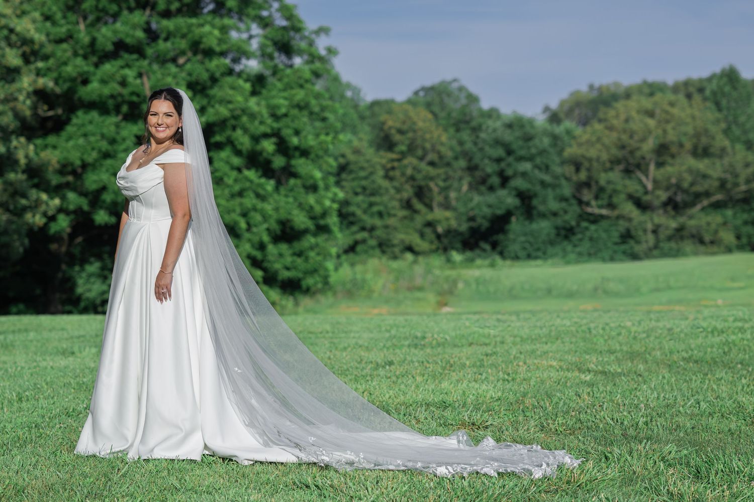 Bride in white gown with long veil standing on green grass field with trees in background.