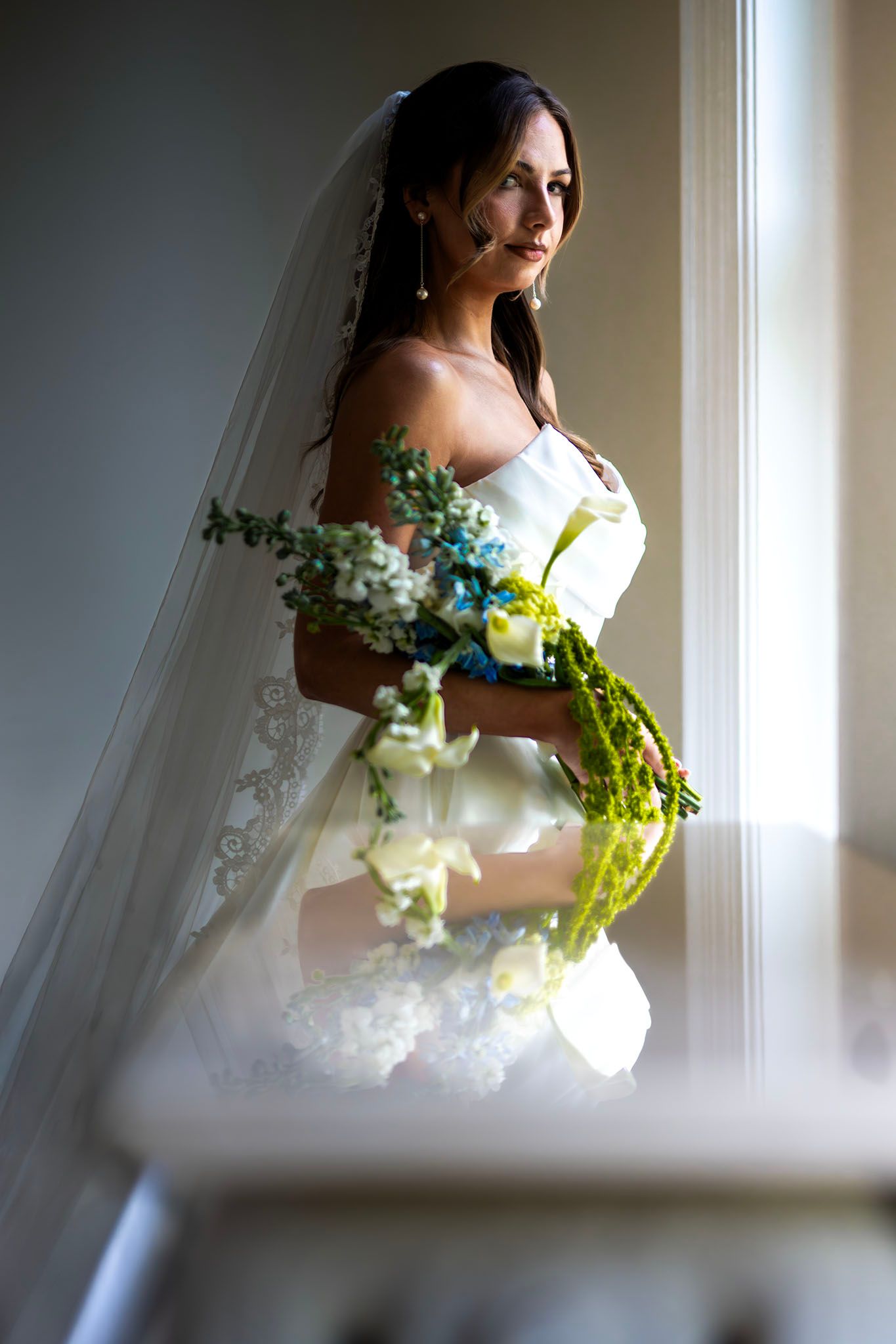 Elegant bride in white off-shoulder gown holding floral bouquet, standing by window light with lace veil.
