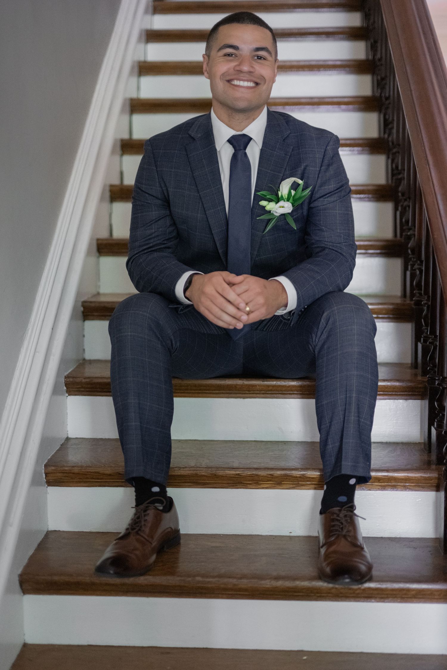 Smiling groom in navy suit with boutonniere sits on wooden staircase, wearing brown dress shoes and dark socks.