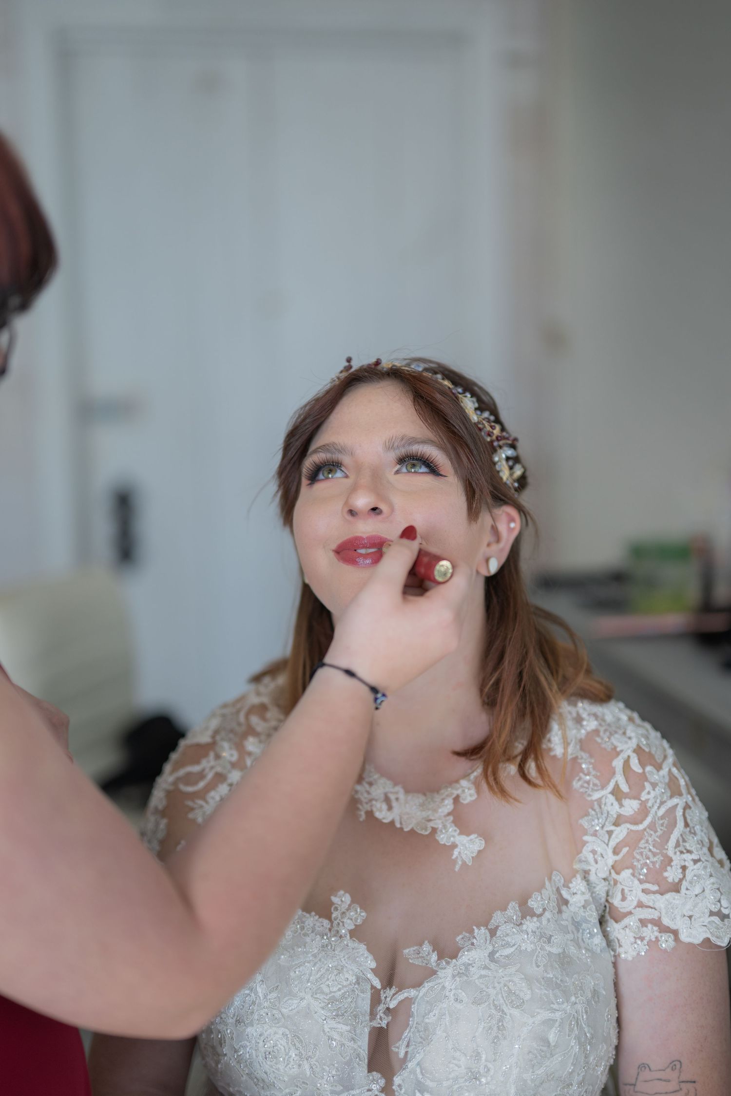 Bride in lace wedding dress getting lipstick applied by makeup artist during bridal preparations.
