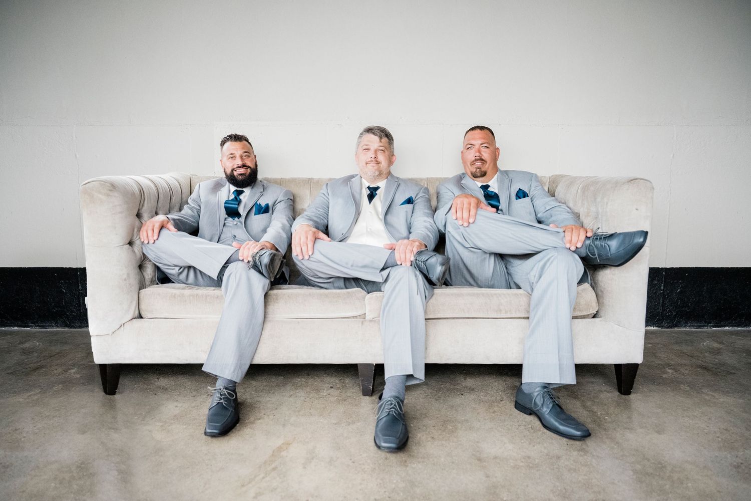 Three groomsmen in light gray suits sitting relaxed on a beige sofa at a wedding venue.