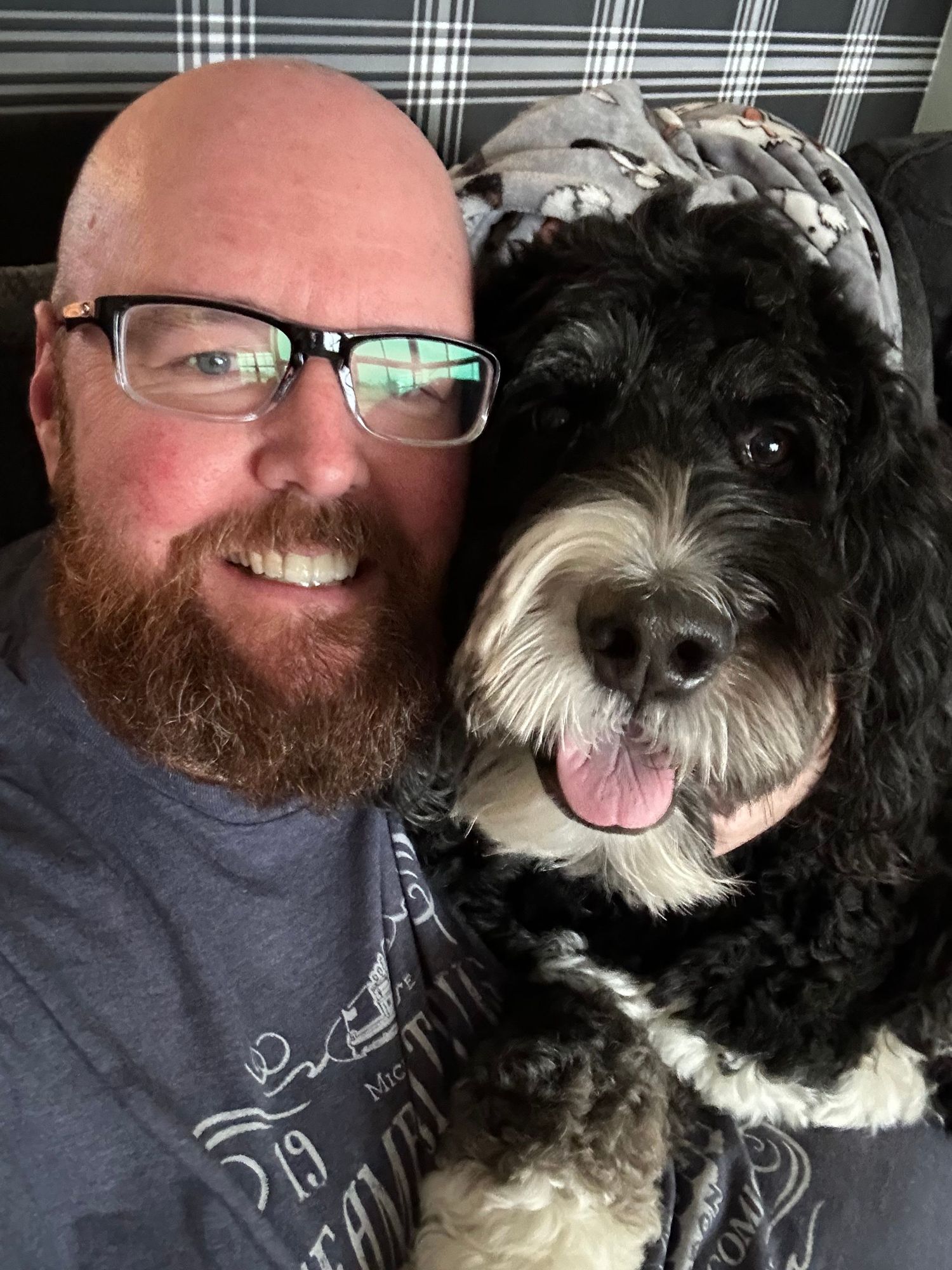 Smiling bald man with red beard and glasses takes selfie with fluffy black and white Bernedoodle dog.
