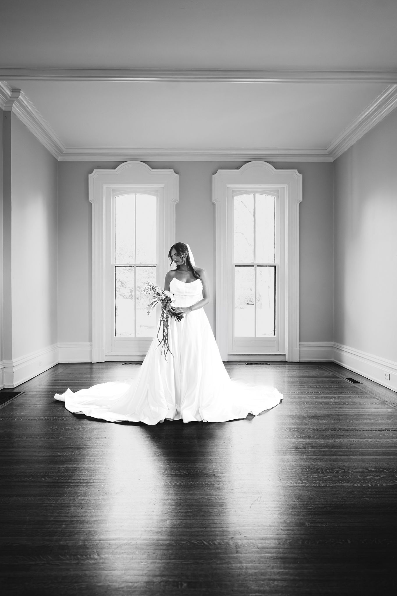 Black and white photo of a bride in a white gown holding a bouquet, standing in an elegant room with hardwood floors.