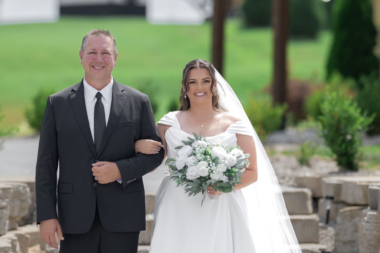 Bride in white gown holding bouquet walks arm-in-arm with man in dark suit at outdoor wedding ceremony.