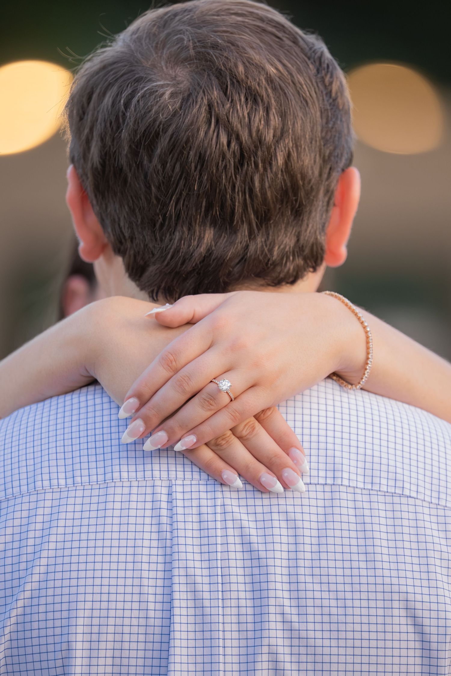 Woman hugging man from behind, displaying diamond engagement ring on her finger, with bokeh lights in background.