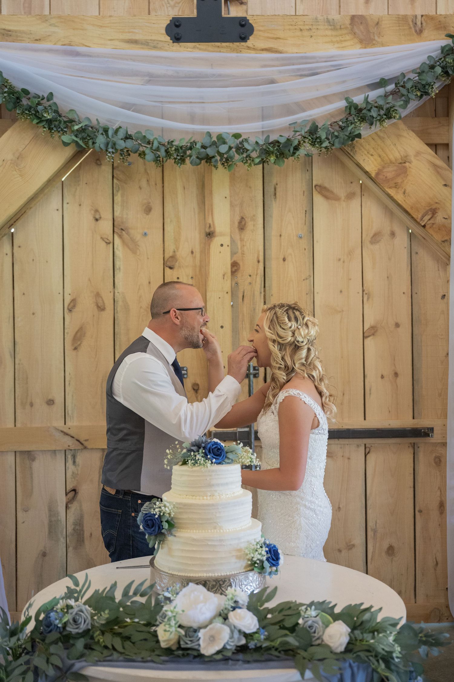 Bride and groom feeding each other wedding cake in front of rustic barn doors with greenery and white floral decor.