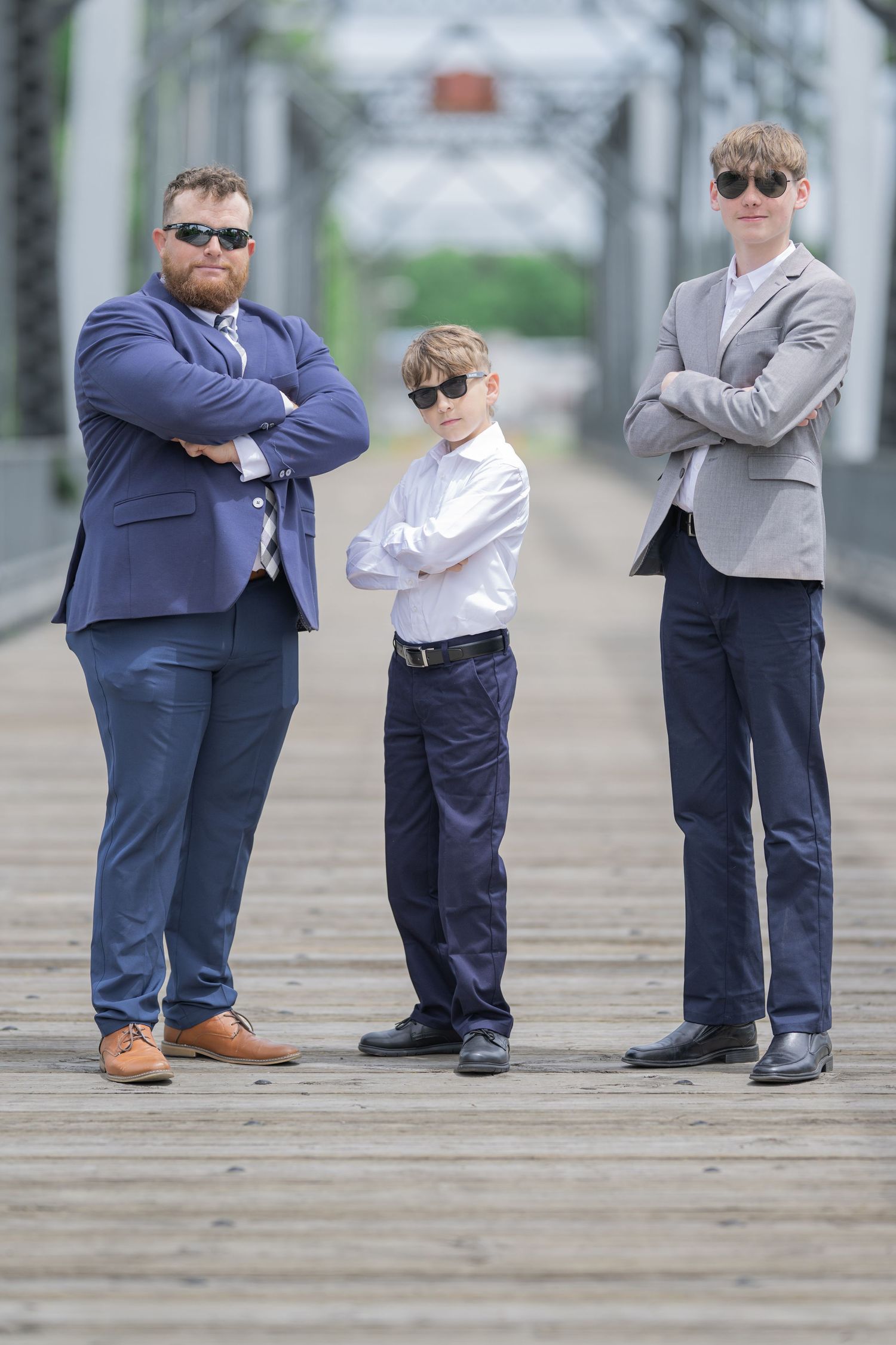 Three males in suits and sunglasses pose confidently with arms crossed on a wooden bridge.