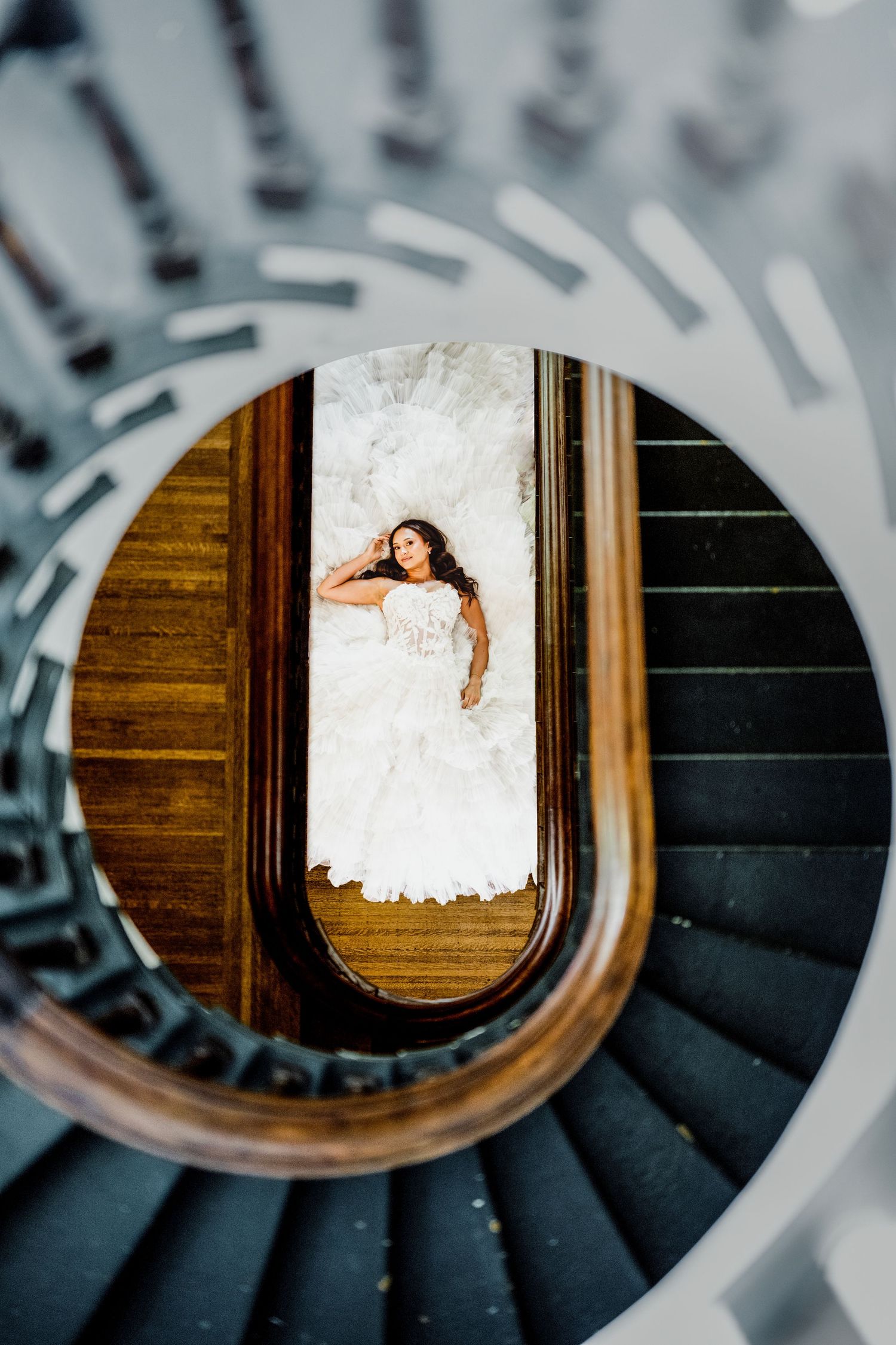 Bride in white wedding gown lying on marble floor, viewed through spiral staircase opening from above.