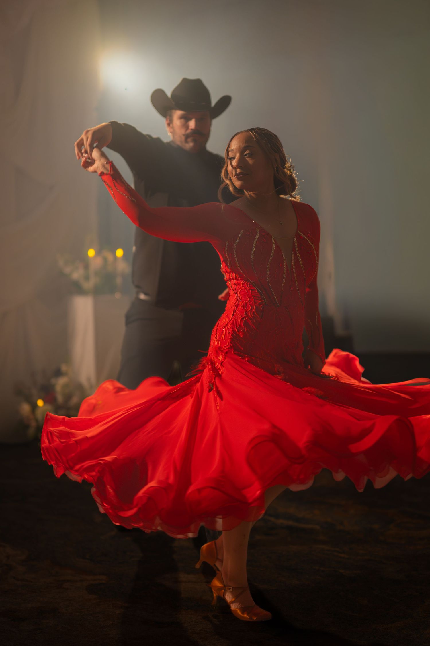 Couple dancing in ballroom, woman in flowing red gown, man in black cowboy hat, dramatic stage lighting.