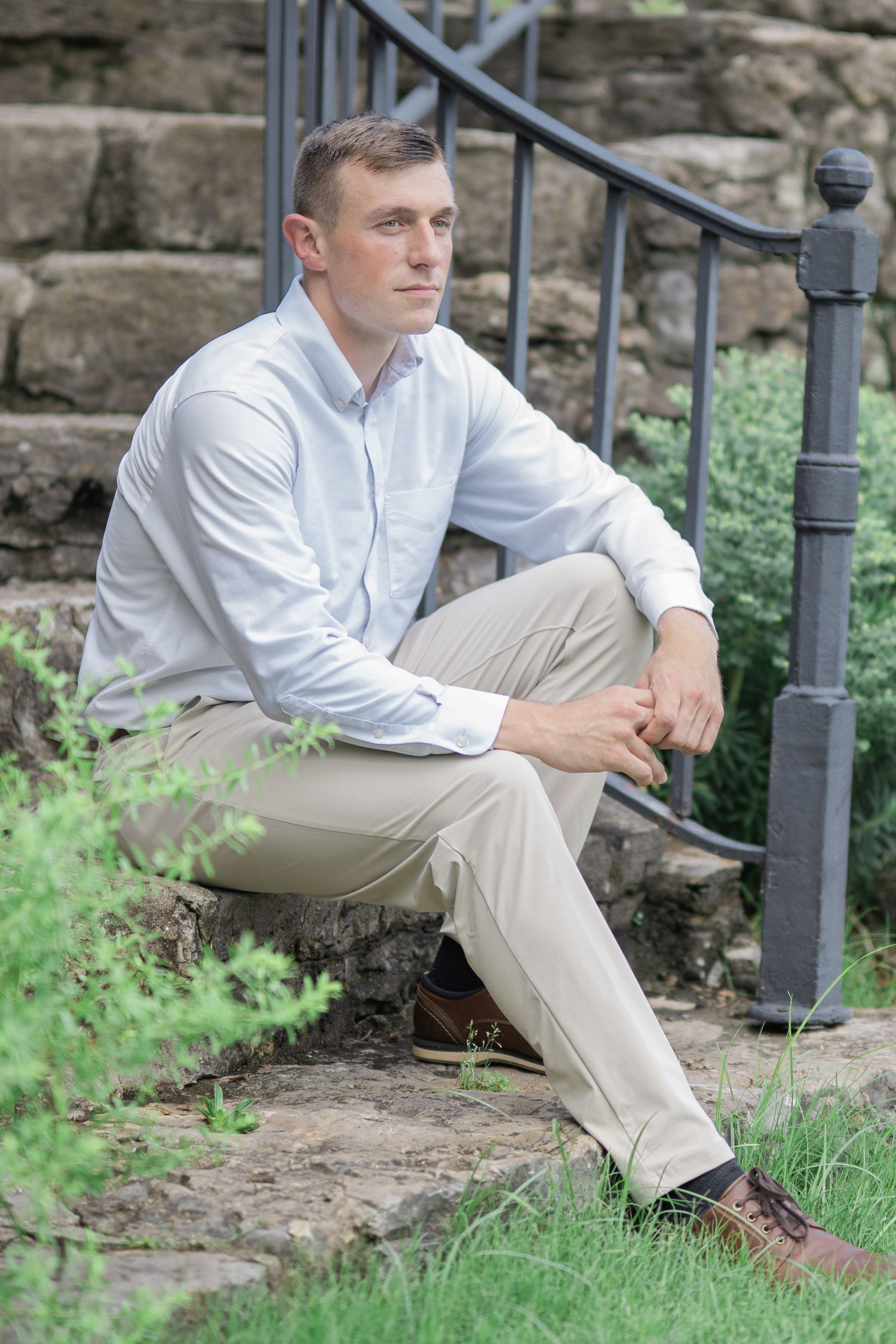 Man in white shirt and khaki pants sitting thoughtfully on stone steps outdoors near green foliage.