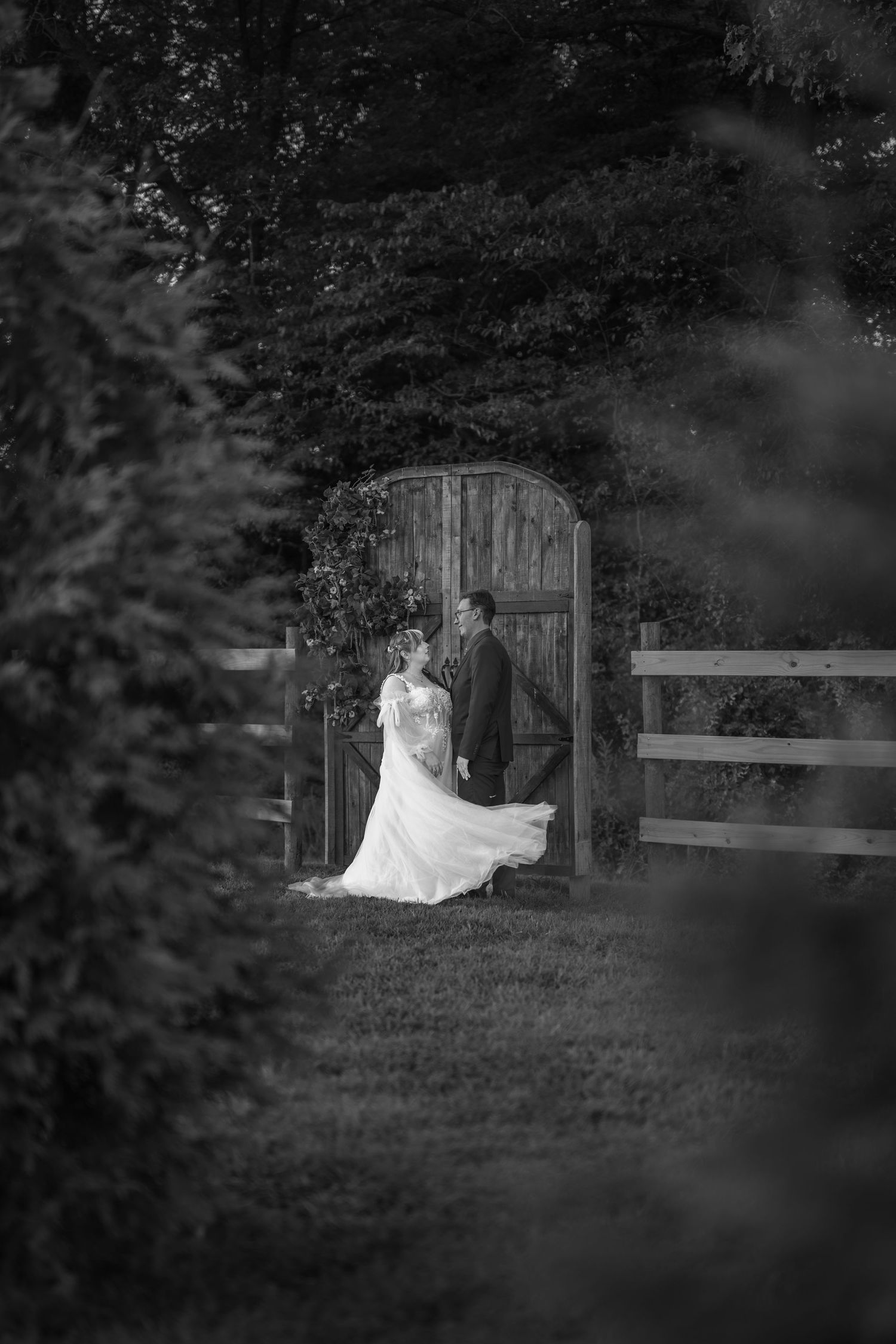 Black and white wedding photo of bride and groom kissing by a rustic wooden gate surrounded by lush greenery.