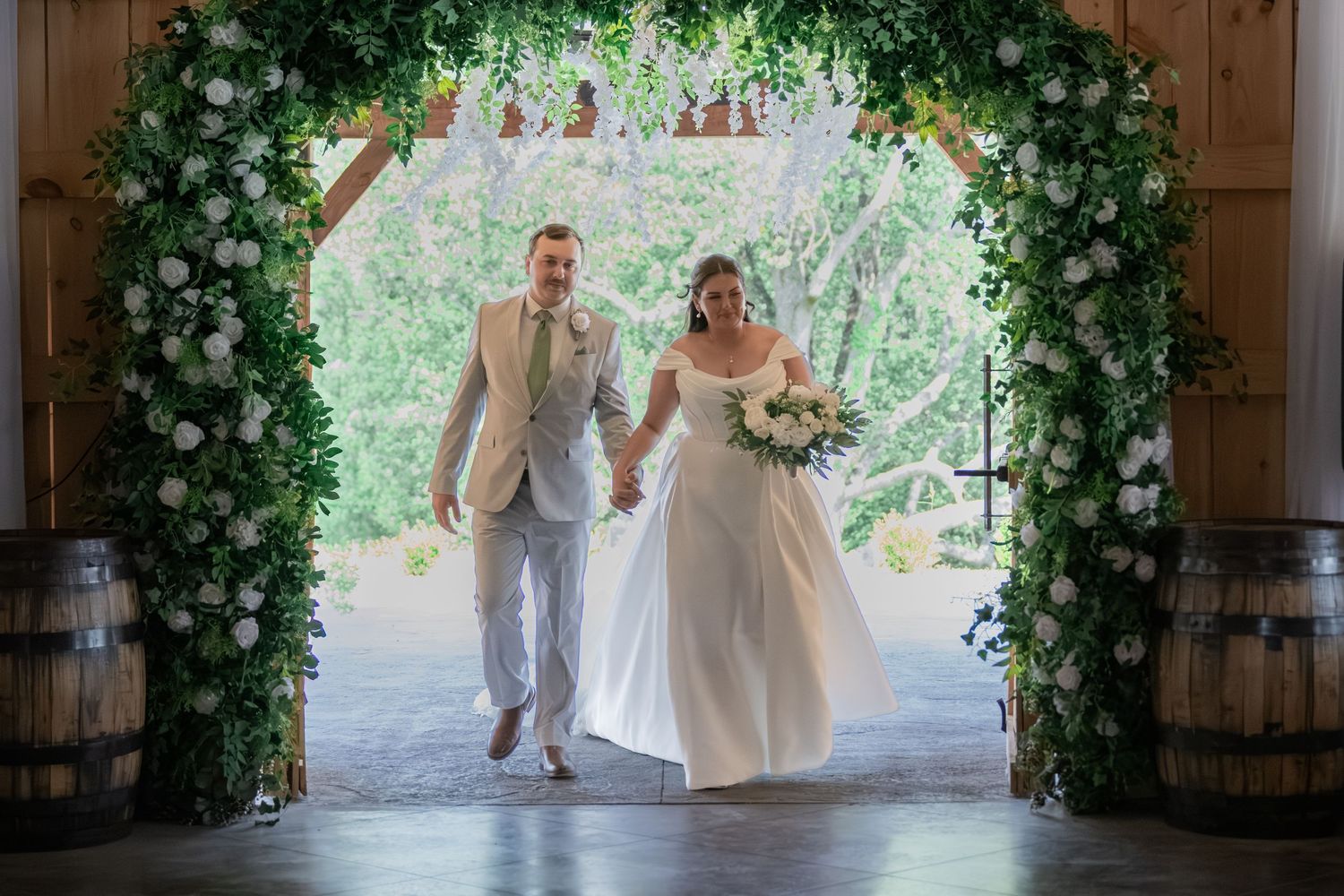 Bride and groom walk hand-in-hand through floral arch of white roses and greenery at rustic barn wedding ceremony.