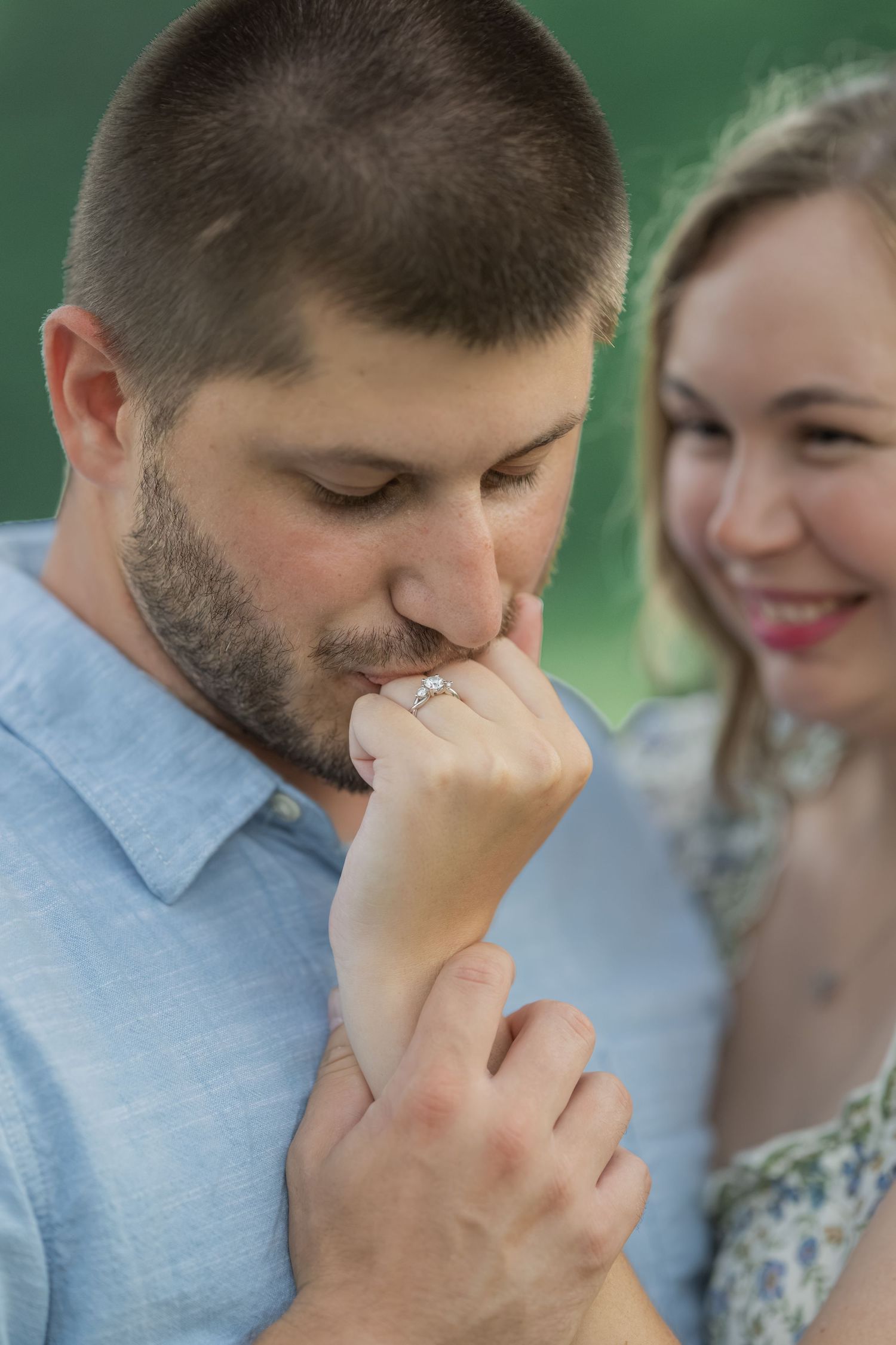 Man kissing woman's hand displaying engagement ring while she smiles joyfully during outdoor proposal moment.