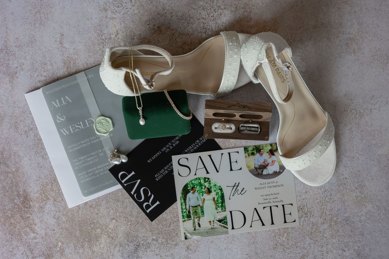White bridal heels beside Save the Date cards, pearl earrings, and a green jewelry box on a marble surface.