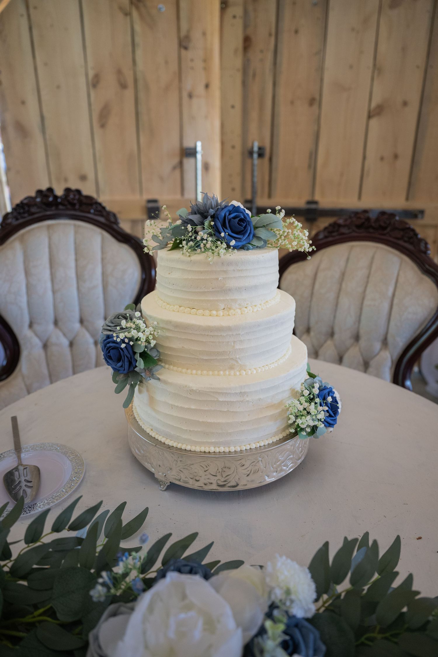 Three-tier white wedding cake with blue roses and greenery on silver stand, displayed between elegant chairs in rustic barn venue.