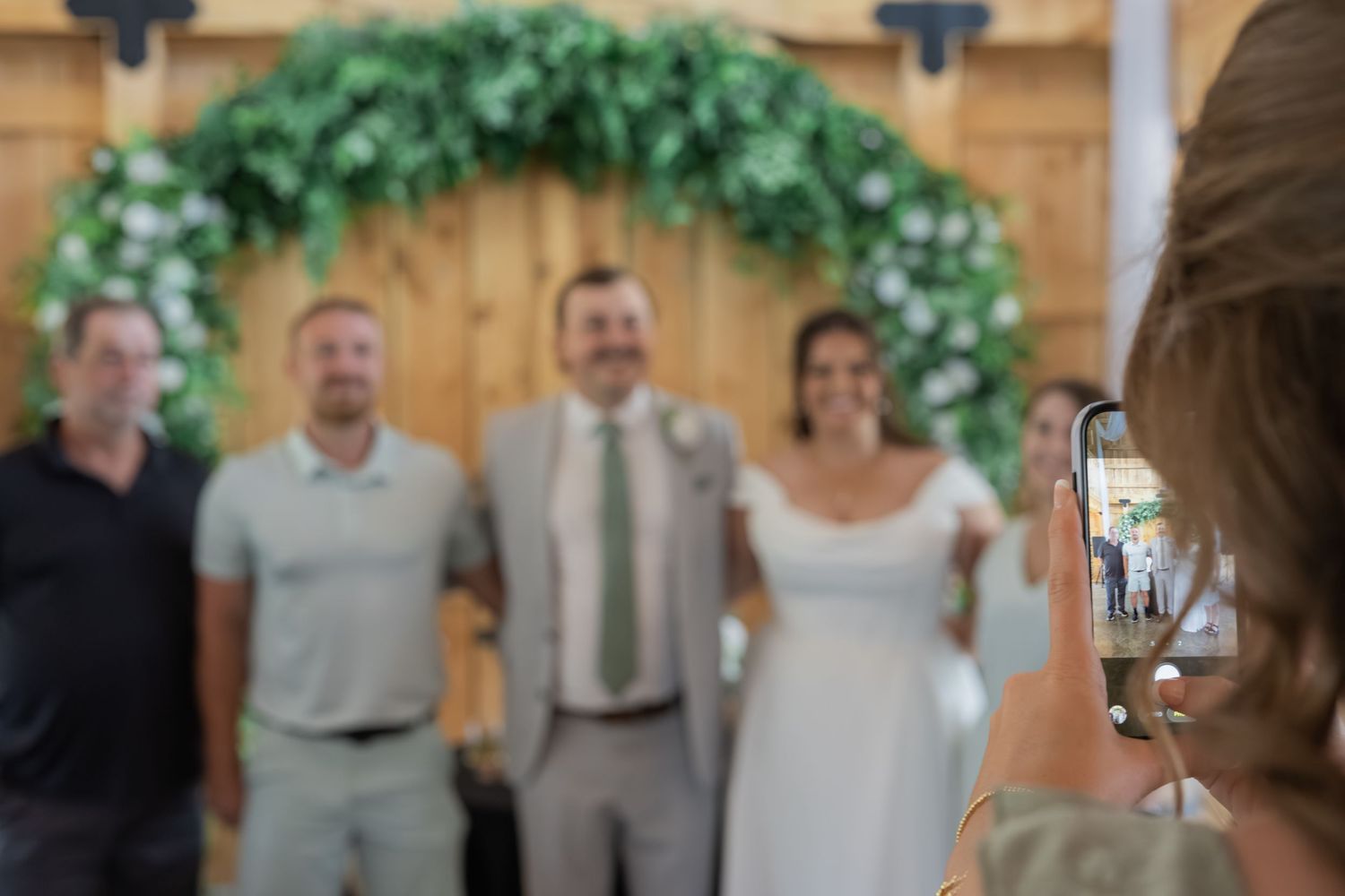 Guest photographs wedding couple with groomsmen under floral arch in rustic barn venue.