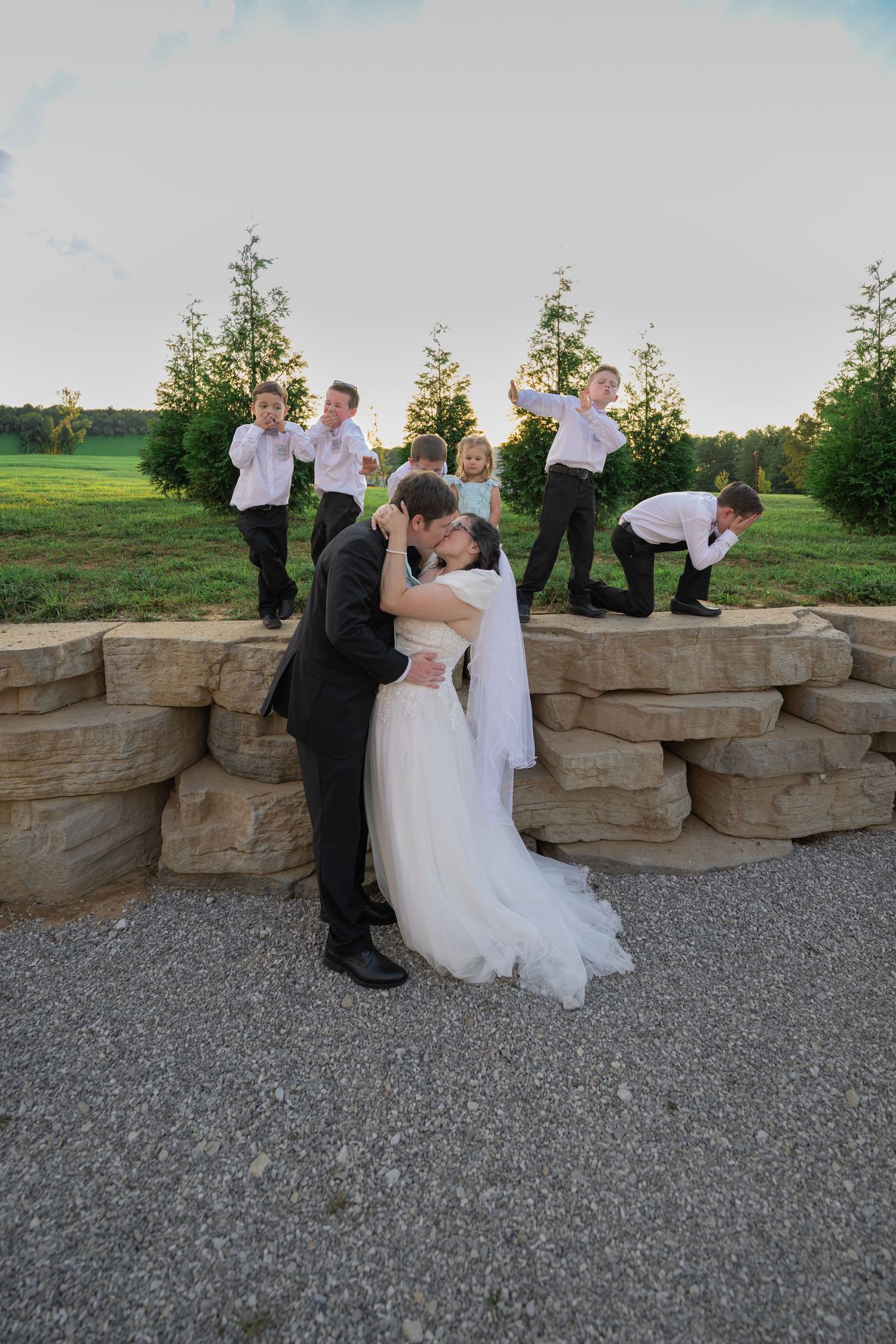 Bride and groom share a kiss outdoors while children play behind a stone wall at a wedding celebration.