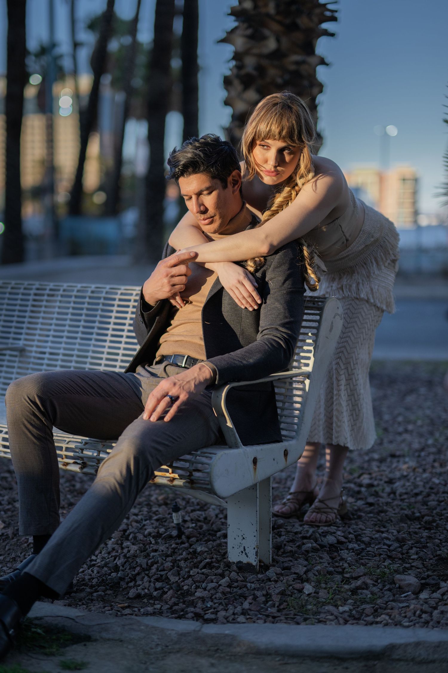 A couple poses on a white bench at dusk, with palm trees and city skyline in the background.