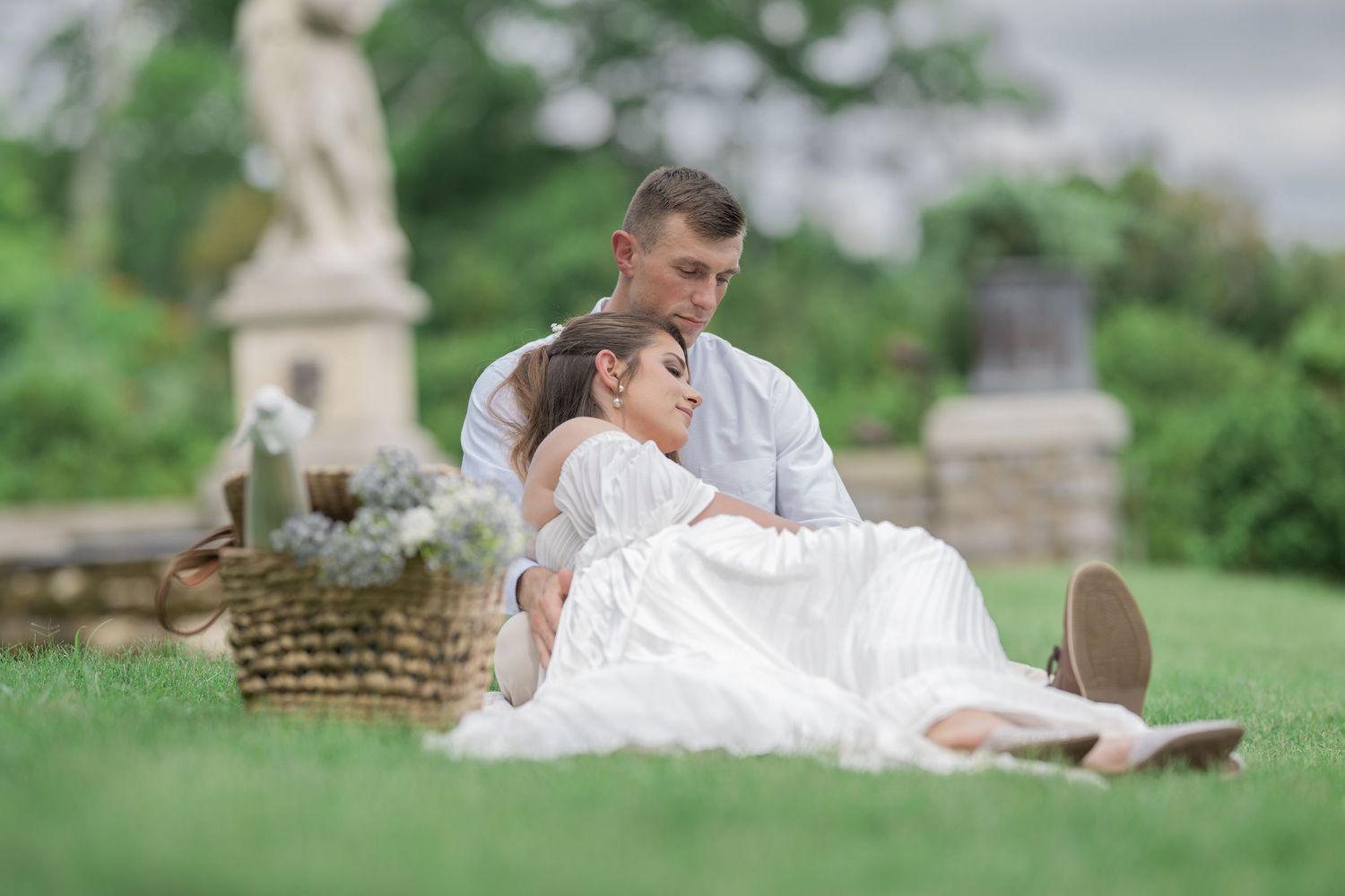 Couple sitting on lush green grass in a garden, woman in white dress leaning against man near a wicker basket.