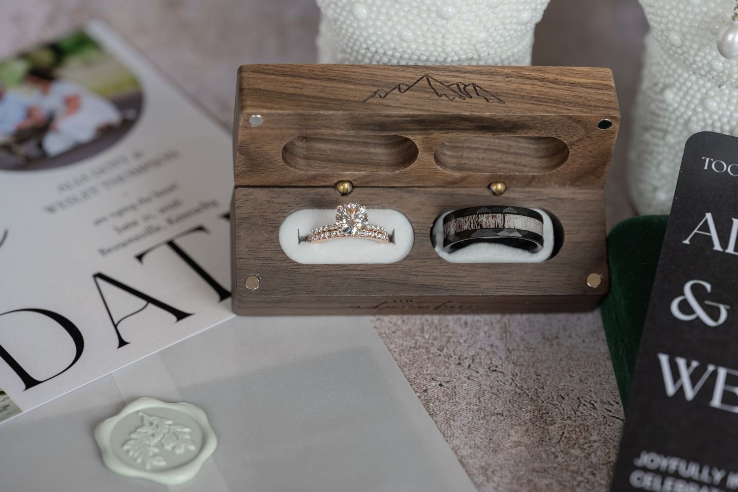 Elegant wooden ring box displaying a diamond engagement ring and black wedding band, placed on wedding stationery with wax seal.