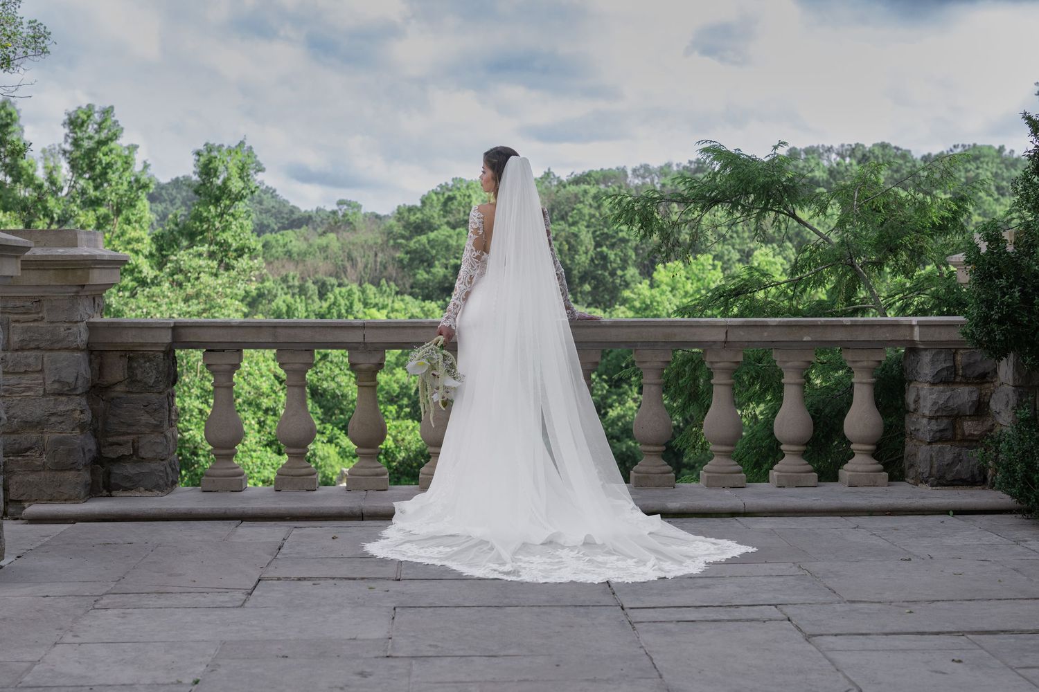 Bride in white wedding gown and long veil stands on stone terrace overlooking lush green garden landscape.