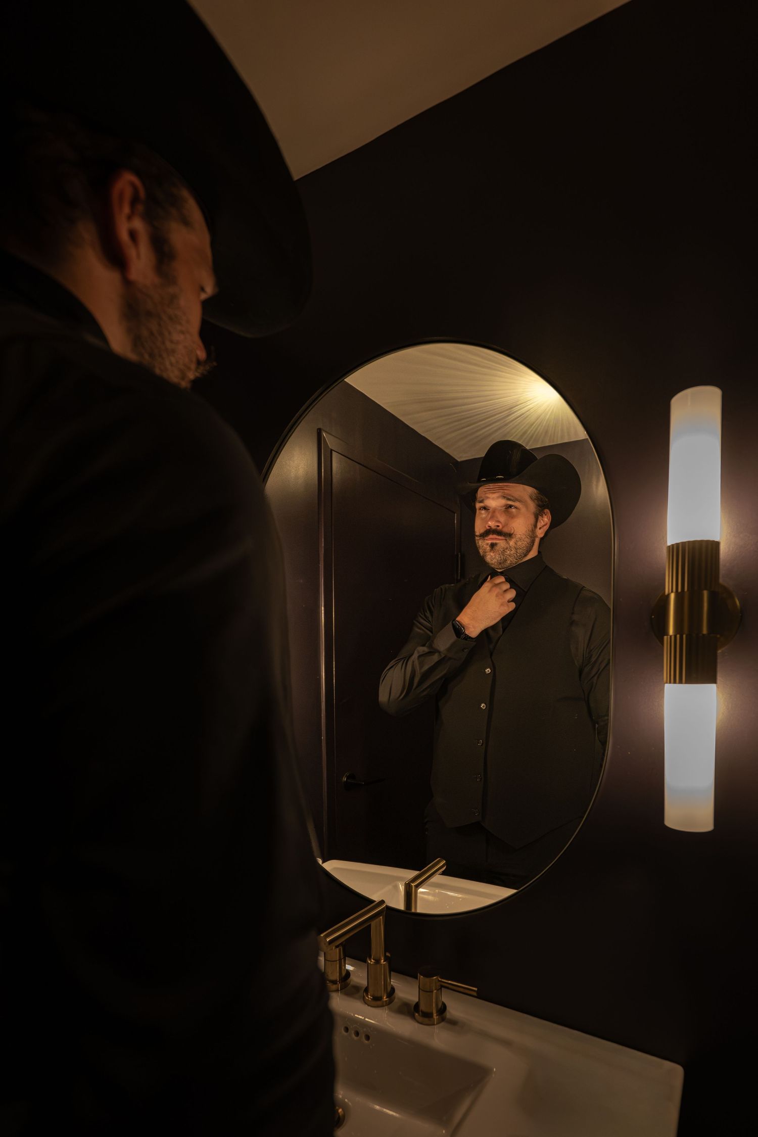 Bearded man in black cowboy hat adjusting collar while smiling at his reflection in a round bathroom mirror.