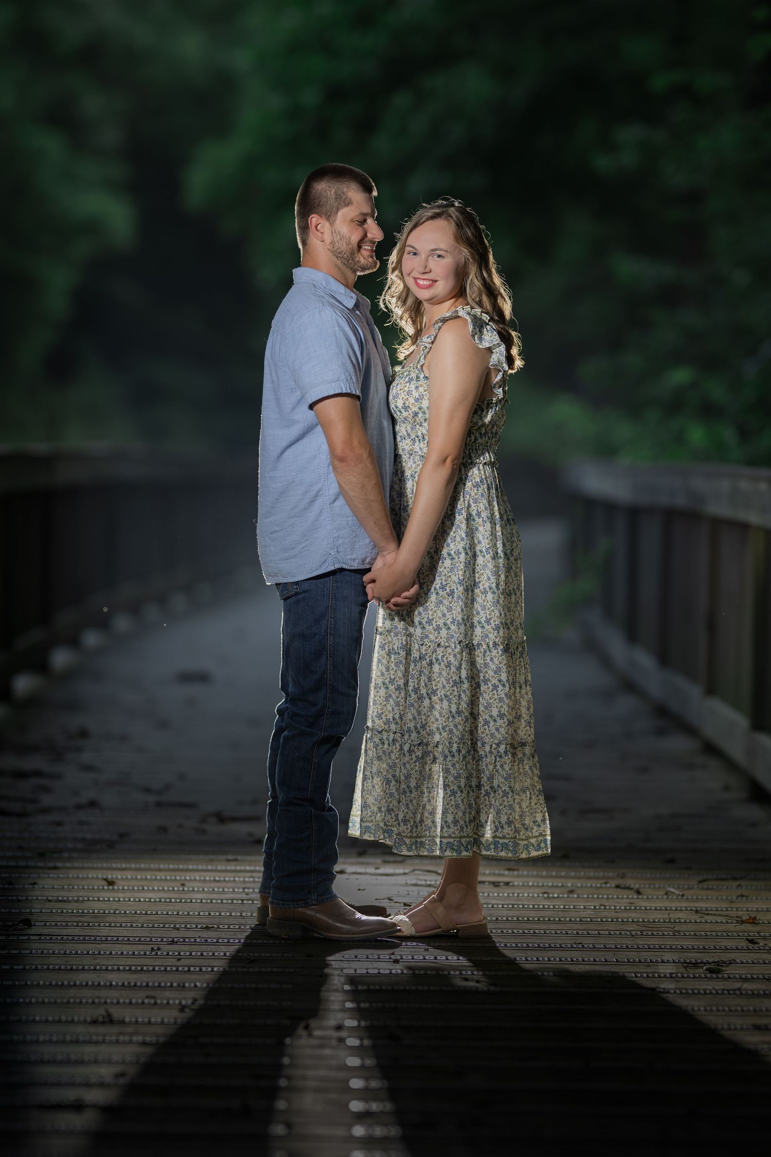 Couple holding hands on a wooden bridge surrounded by lush green trees, woman in floral dress smiling at camera.