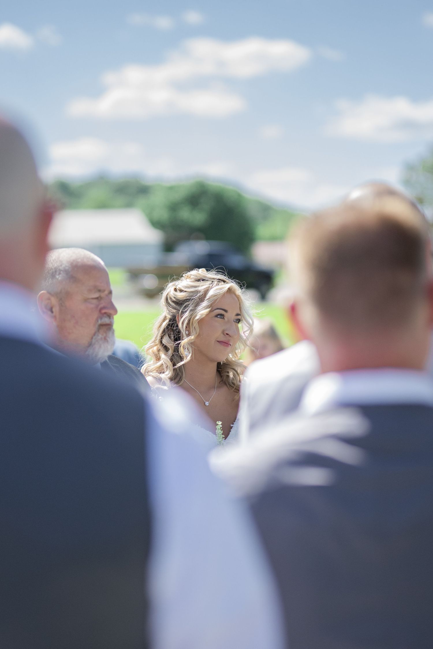 Bride with curly blonde hair stands outdoors at a sunny wedding ceremony surrounded by guests in formal attire.