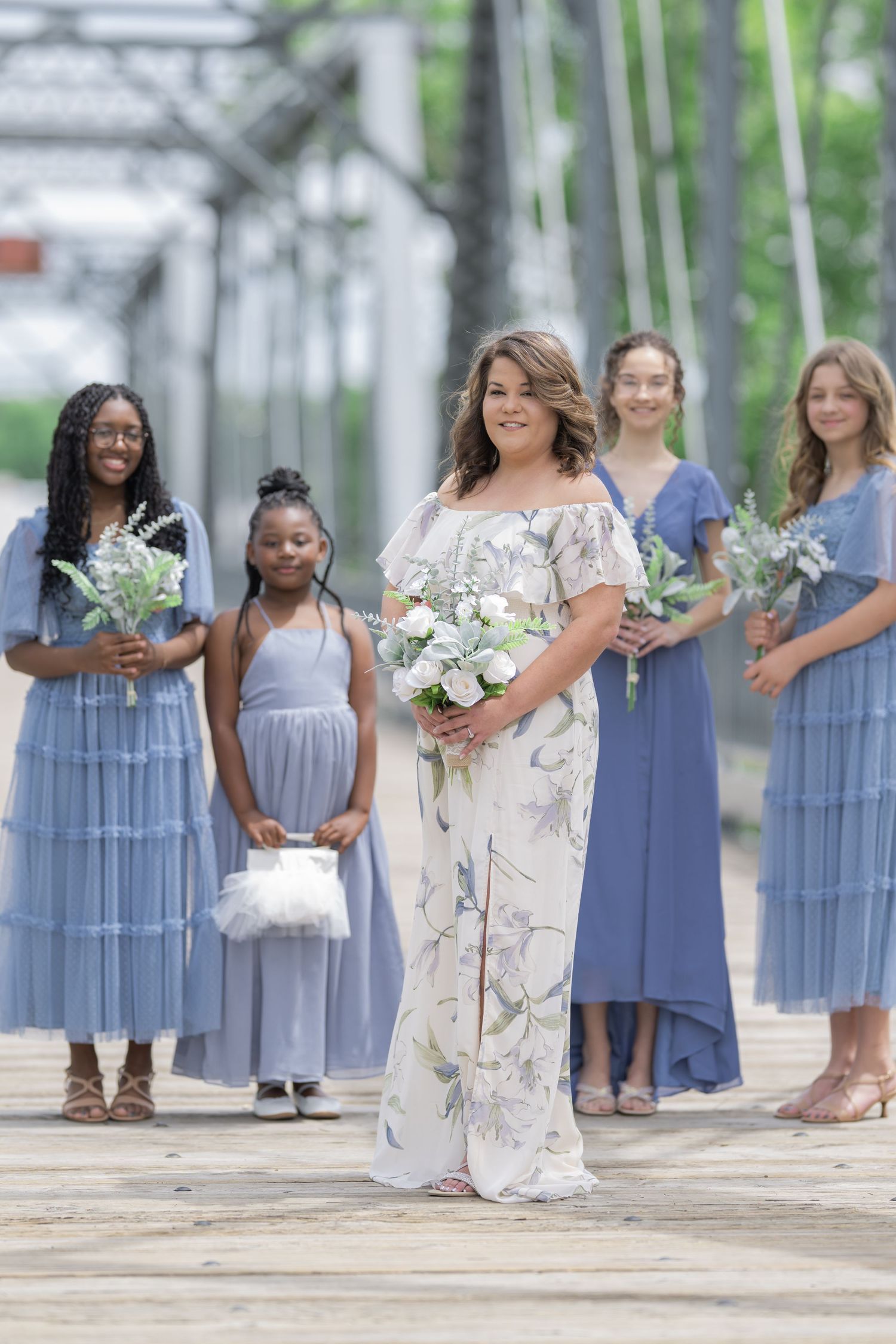 Bride in floral gown holds white bouquet with bridesmaids in blue dresses on wooden bridge.