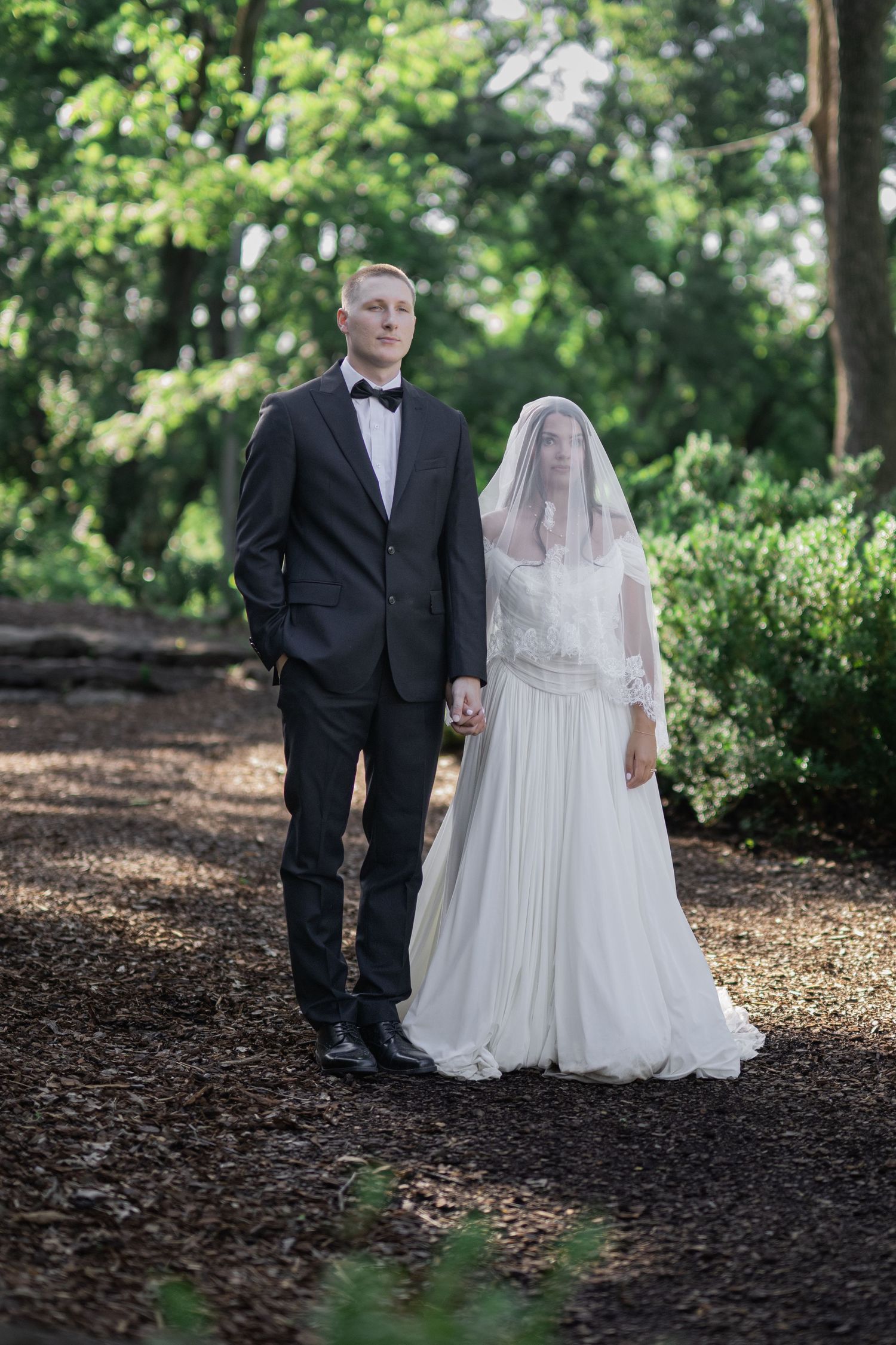 Bride and groom holding hands on a woodland path, she wears a white gown with veil, he wears a black tuxedo.