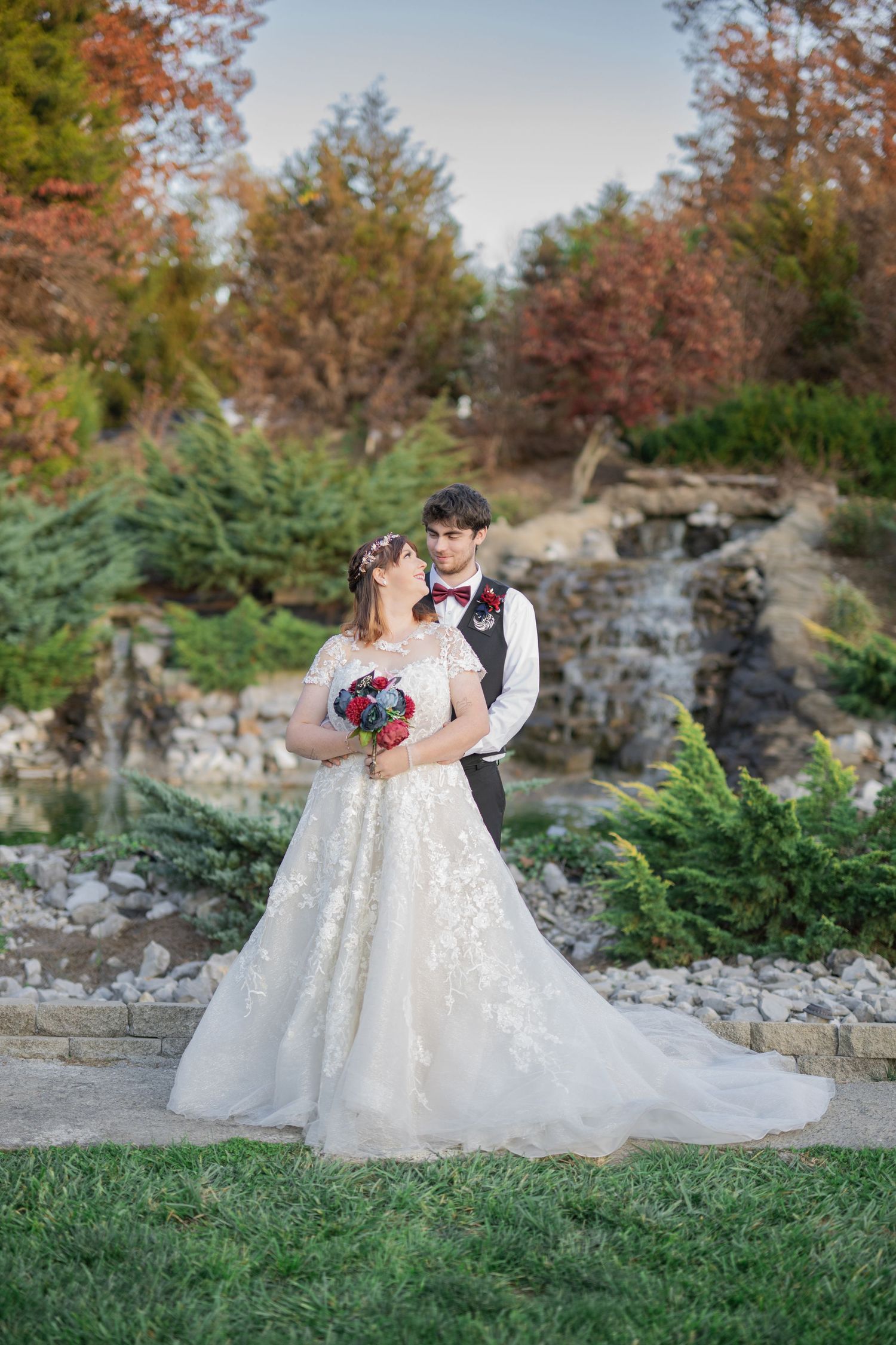 Bride and groom embrace in a lush garden with a waterfall backdrop during autumn wedding photo session.