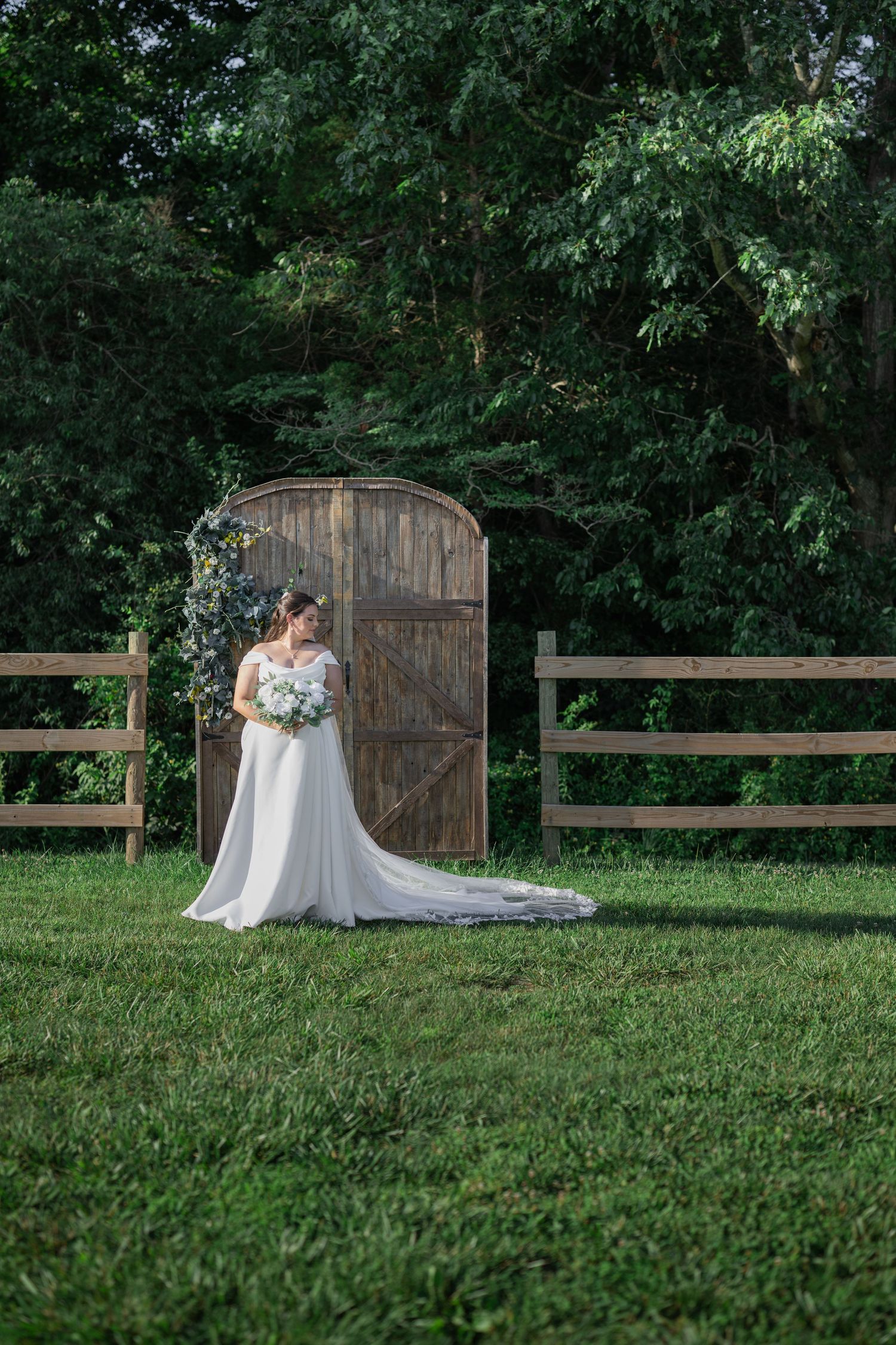 Bride in white gown holding bouquet stands before rustic wooden gate surrounded by lush greenery on a grassy lawn.