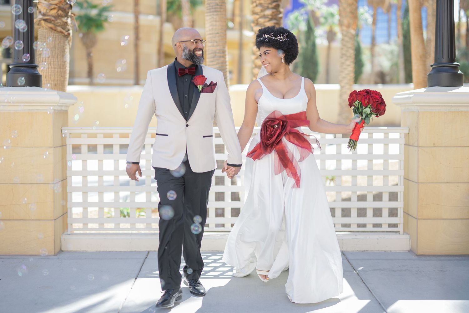 Bride and groom walk together outdoors, she in white gown with red sash, he in white tuxedo with red boutonniere.