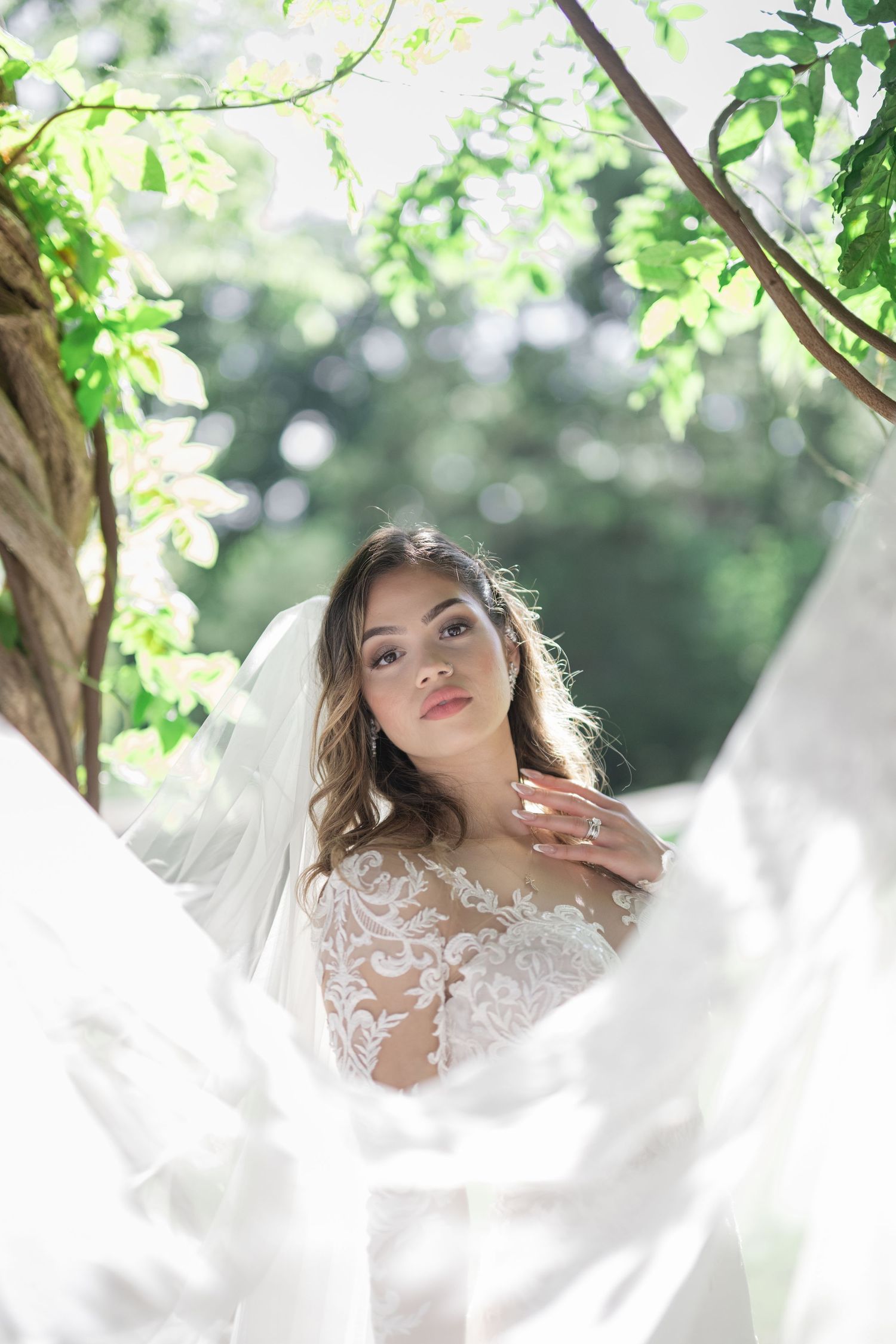 Elegant bride in lace wedding dress and veil poses under green tree canopy in a sunlit outdoor garden setting.