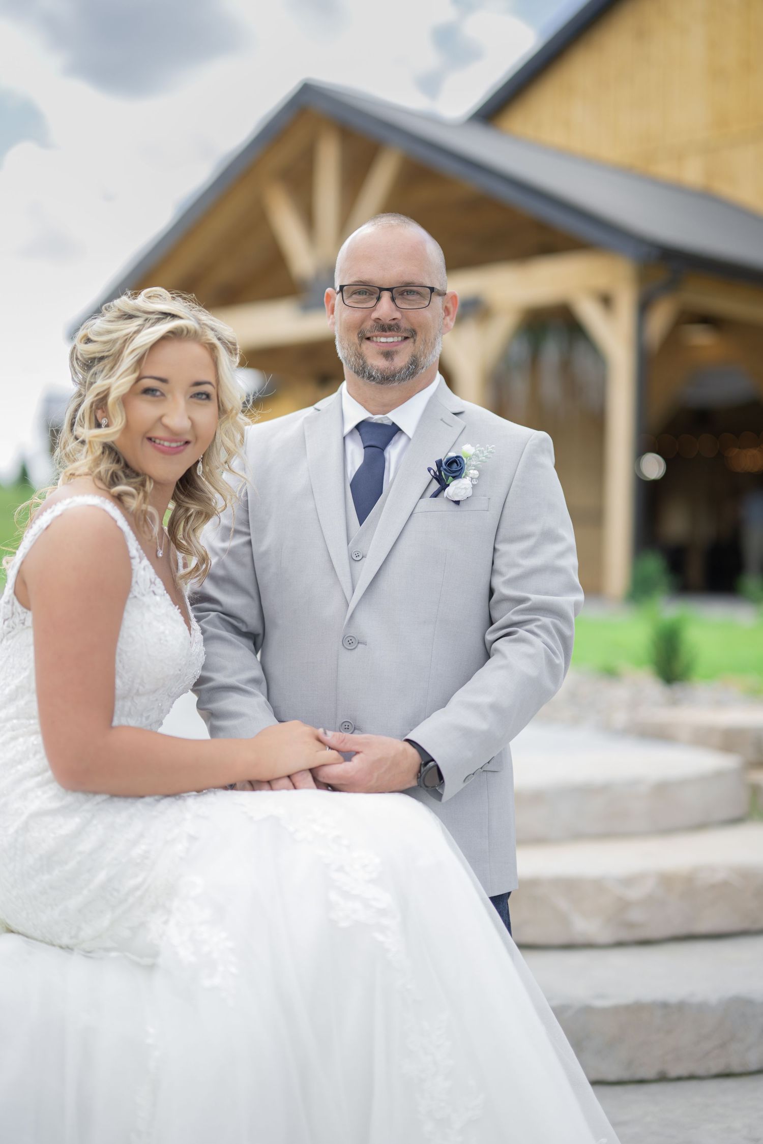 Smiling bride and groom pose together on stone steps outside a wooden venue on their wedding day.