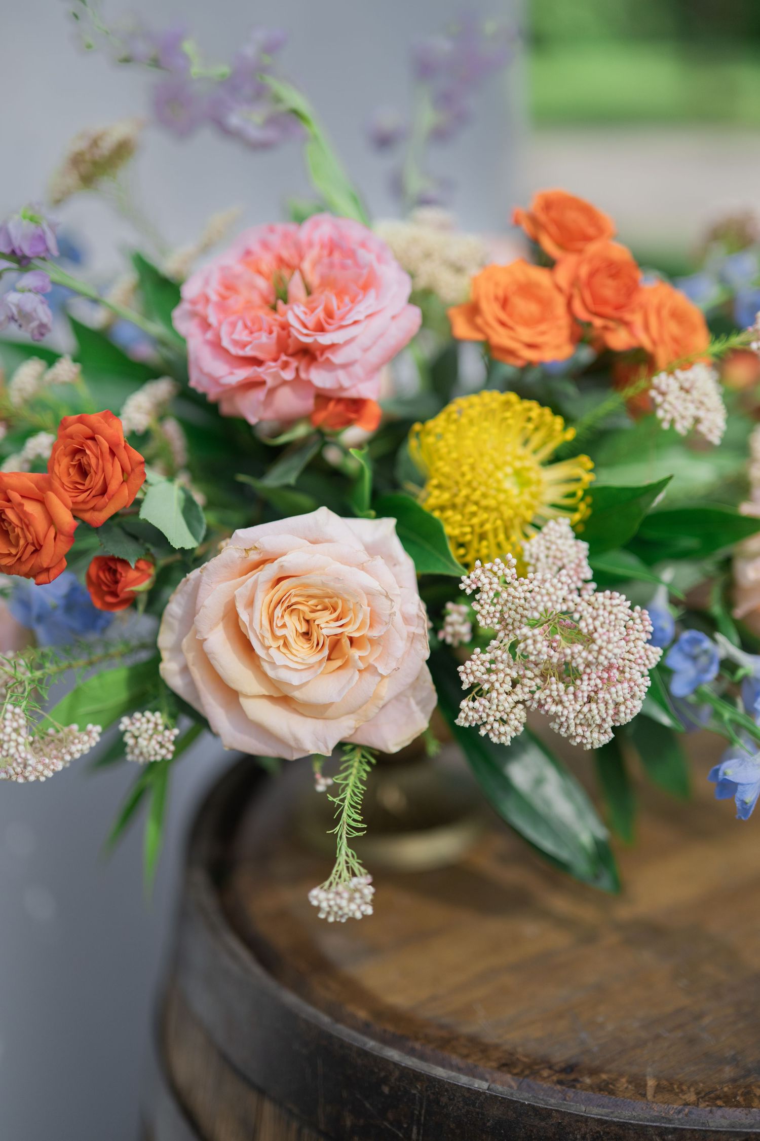 Colorful floral arrangement with peach, orange, pink roses, yellow chrysanthemum, and blue flowers on a rustic wooden barrel.