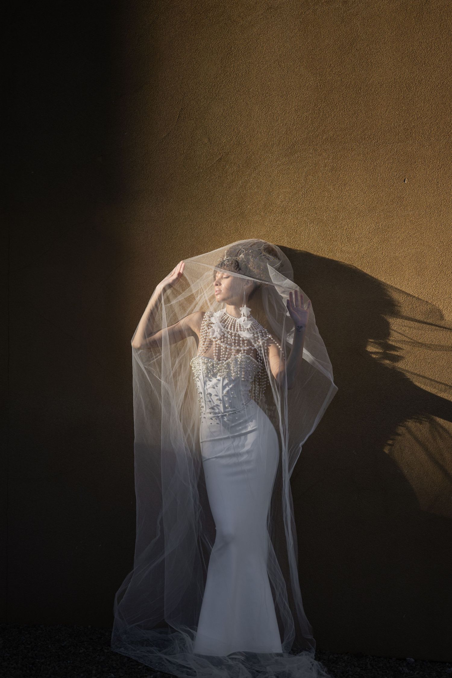 Bride in white mermaid gown and sheer veil posed dramatically against golden wall in artistic light.